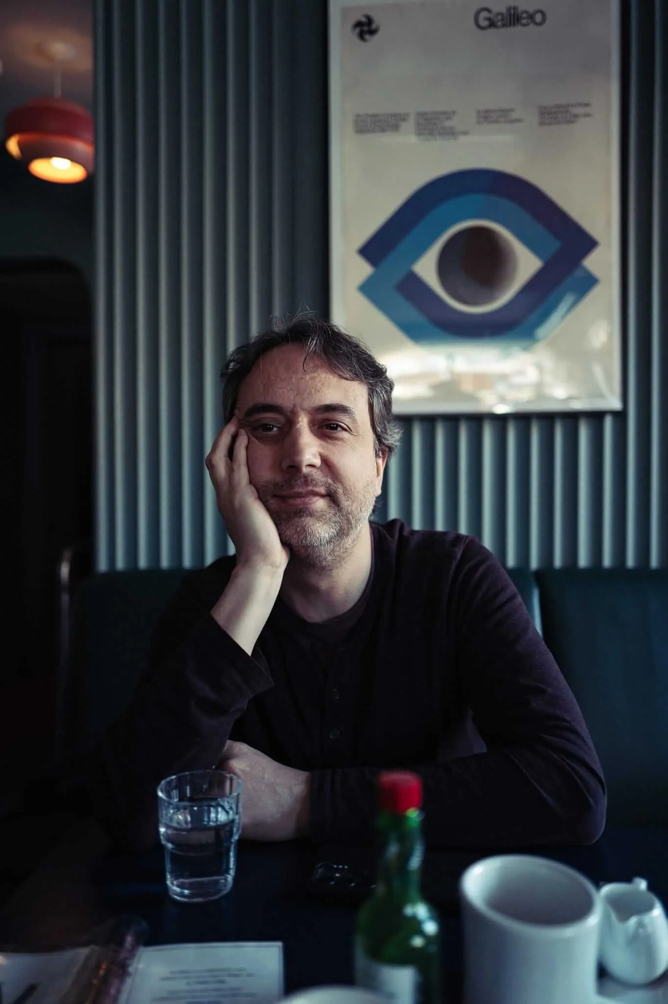 A contemplative middle-aged man with beard and gray hair rests his chin on his hand while sitting at a café table, with a glass of water, coffee cup, and bottle visible. Behind him is a white poster featuring a stylized blue eye logo for 'Galileo' against dark vertical blinds, with warm lighting creating an intimate atmosphere.