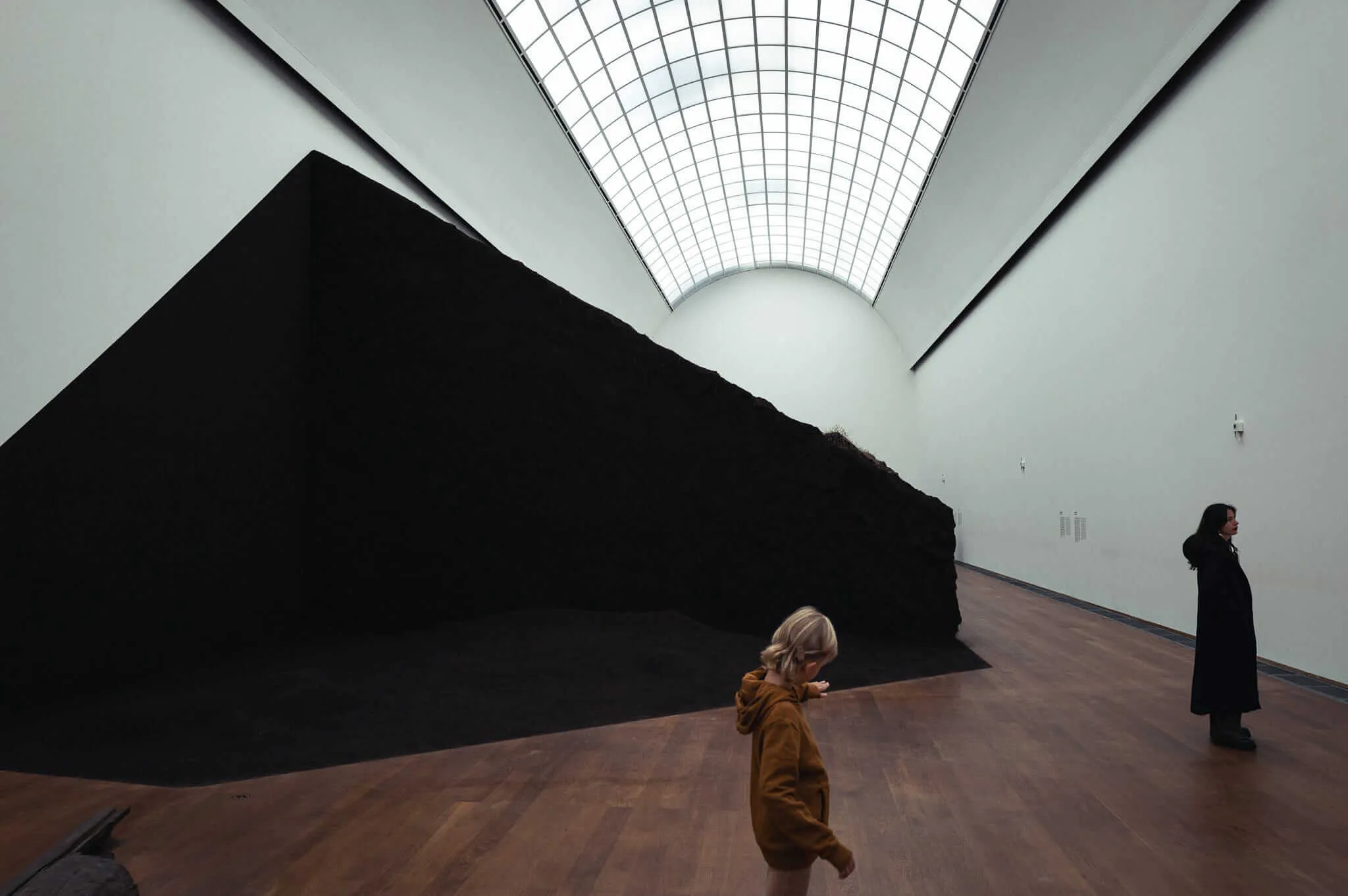 Interior of a modern art gallery with a dramatic large black sculptural form resembling a mountain or angular mass dominating the space. Two visitors in coats observe the installation - one person in brown/tan clothing in the foreground and another in black clothing in the background. The room features white walls, polished wooden floors, and a distinctive curved glass ceiling with geometric grid patterns that allows natural light to filter in from above.