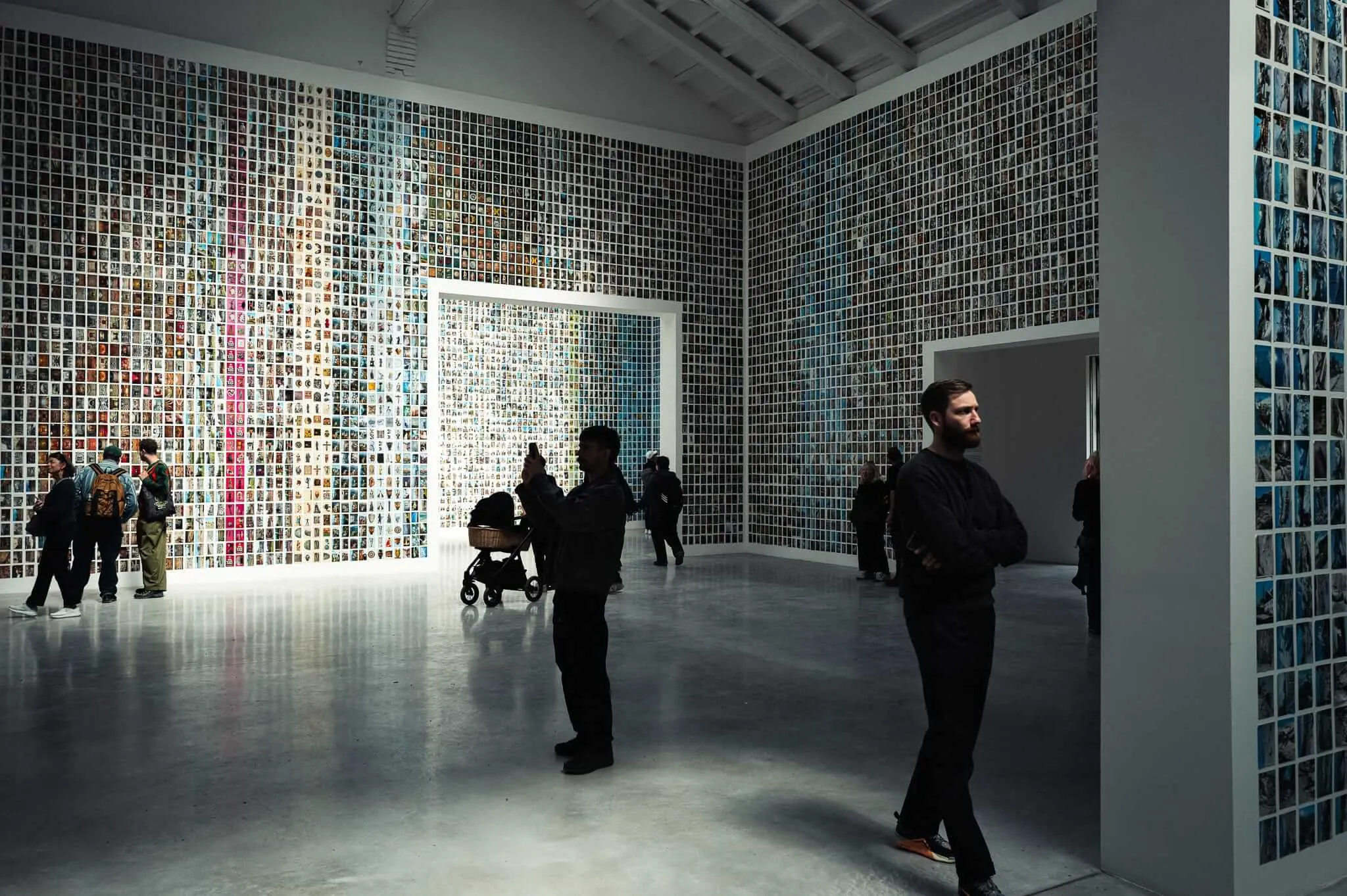 Visitors stand in a large contemporary gallery space whose floor-to-ceiling walls are completely covered in a dense grid of thousands of small images. A bearded man in black stands in the foreground with arms crossed, while a silhouetted figure raises a smartphone to photograph the installation. Further back, visitors with a pram explore the space, and additional rooms visible through doorways reveal more image-covered walls.