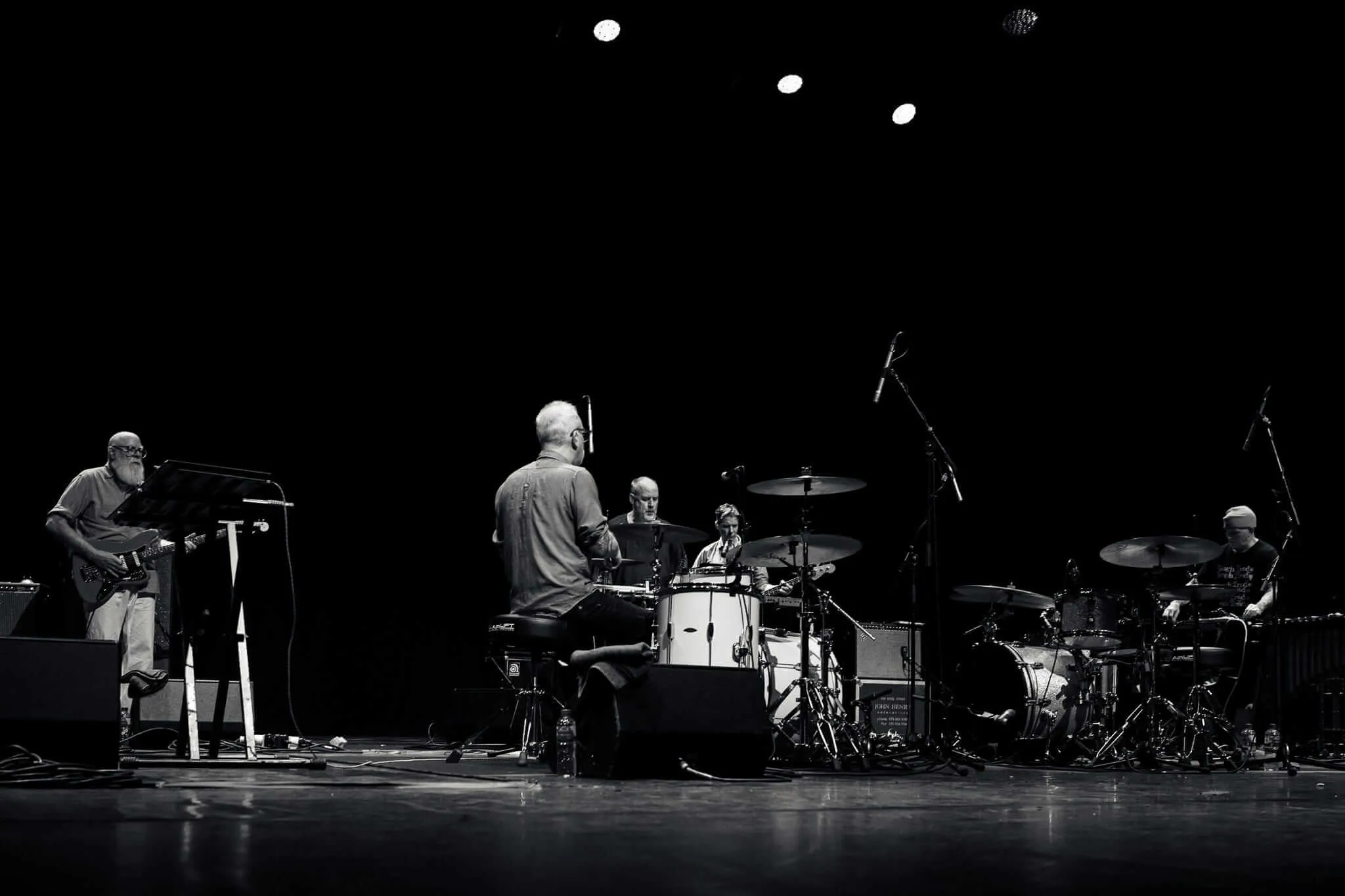 Black and white photograph of five musicians performing on a dark stage with dramatic overhead lighting. From left to right: a guitarist, a musician with his back to the audience, a musician at an electronic kit, another musician at another electronic kit, and a musician on the guitar. The stage floor is reflective and the background is completely black, creating a stark, intimate concert atmosphere.