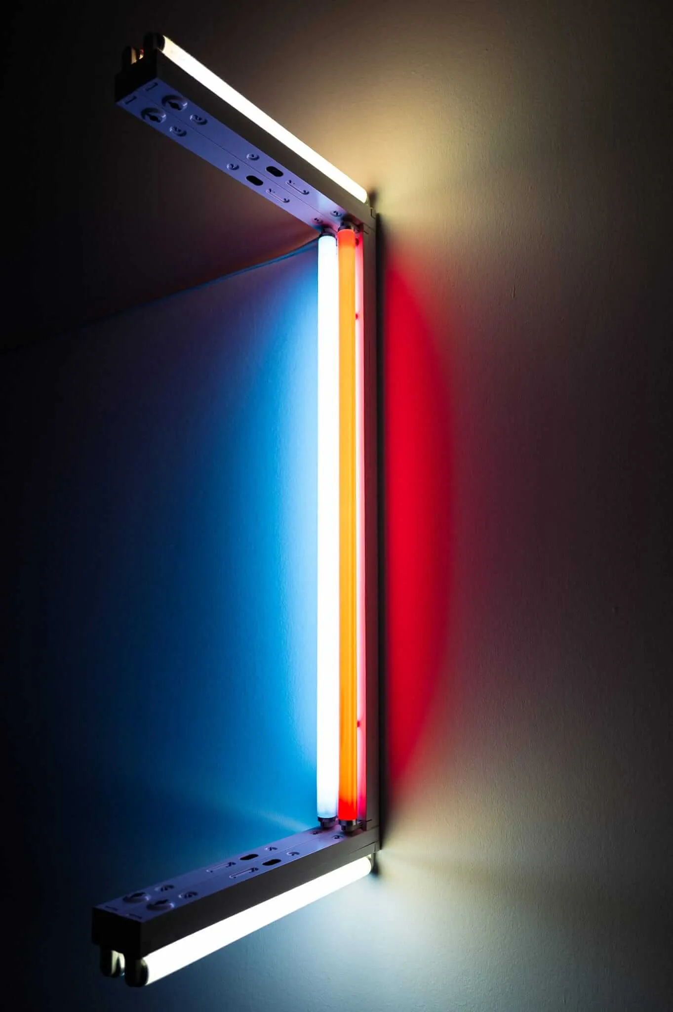 A wall-mounted light installation featuring vertical fluorescent tubes in blue, white, yellow, and red colors creating dramatic colored light beams against a dark wall, with visible mounting hardware and power outlets
