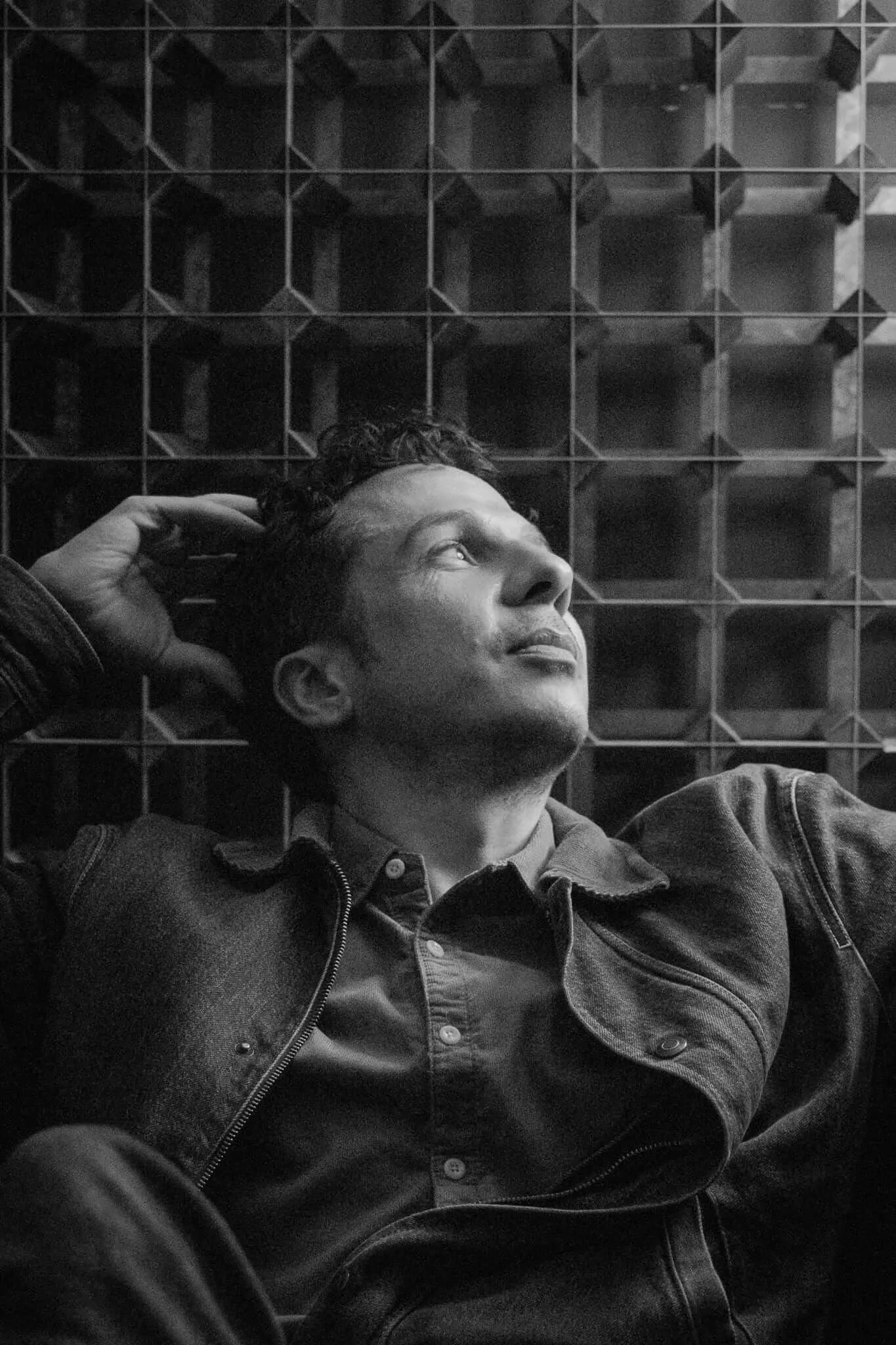 A man in a denim jacket reclines and gazes upward with one hand resting behind his head, set against a geometric brutalist grid structure, in a moody black and white portrait.