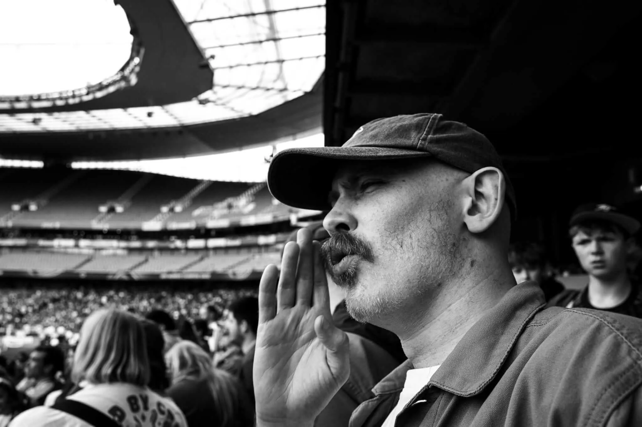 A black and white photograph of a man in a baseball cap cupping his hand around his mouth to amplify his voice while shouting at a football stadium. He has a mustache and goatee, and other spectators are visible in the blurred background. The stadium's distinctive architecture with its curved roof and multiple tiers is visible.
