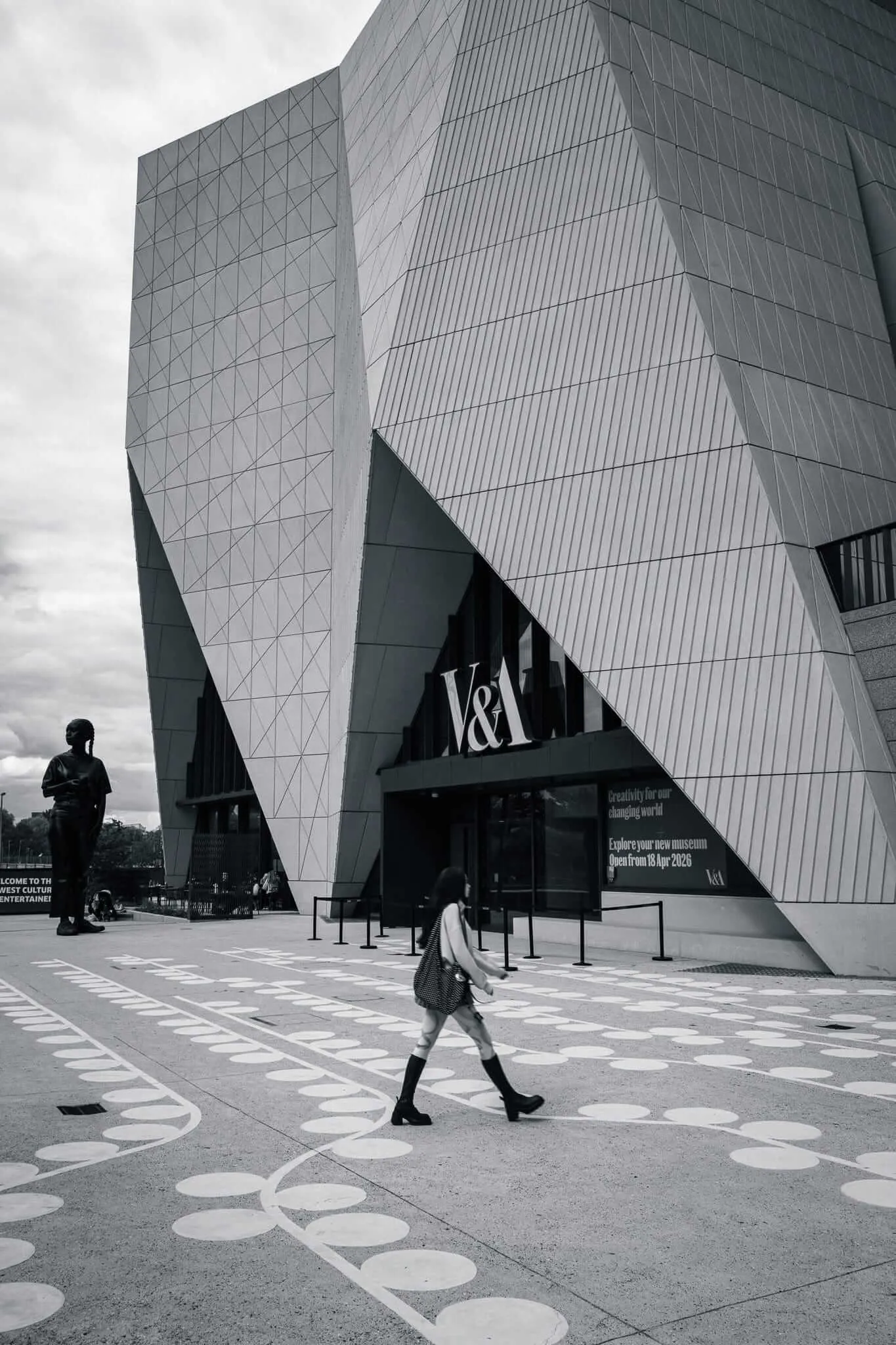 Black and white photograph showing a person walking past the striking geometric facade of the V&A East Museum, with angular metallic cladding creating dramatic shadows and patterns. White circular markers are embedded in the ground forming a pathway, while the building's modern architecture features sharp triangular forms and a bold V&A sign.