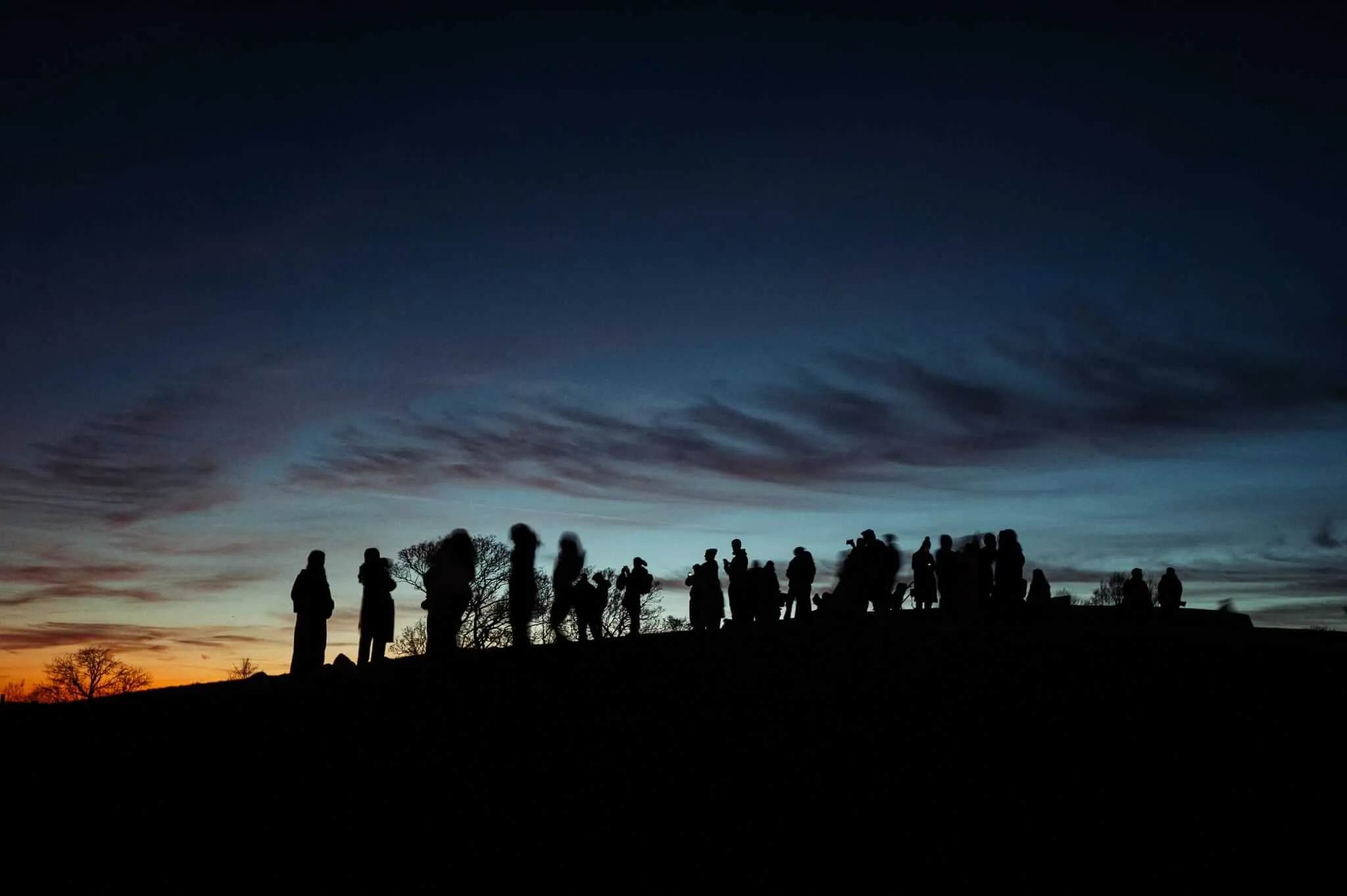 Silhouettes of a large group of people standing on a hillside against a dramatic twilight sky with deep blue clouds above and warm orange light near the horizon. Bare trees frame the scene as the crowd appears to be gathered for an event or gathering during the golden hour.
