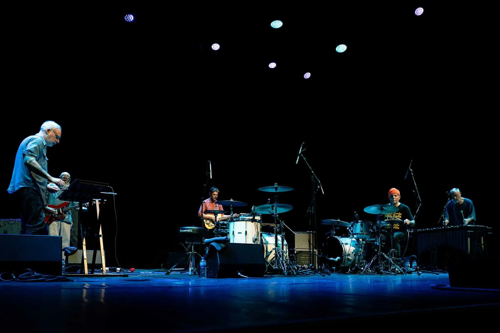 A band performs on a dark stage illuminated by blue stage lights, with five musicians. Circular stage lights create a starfield effect against the black backdrop.