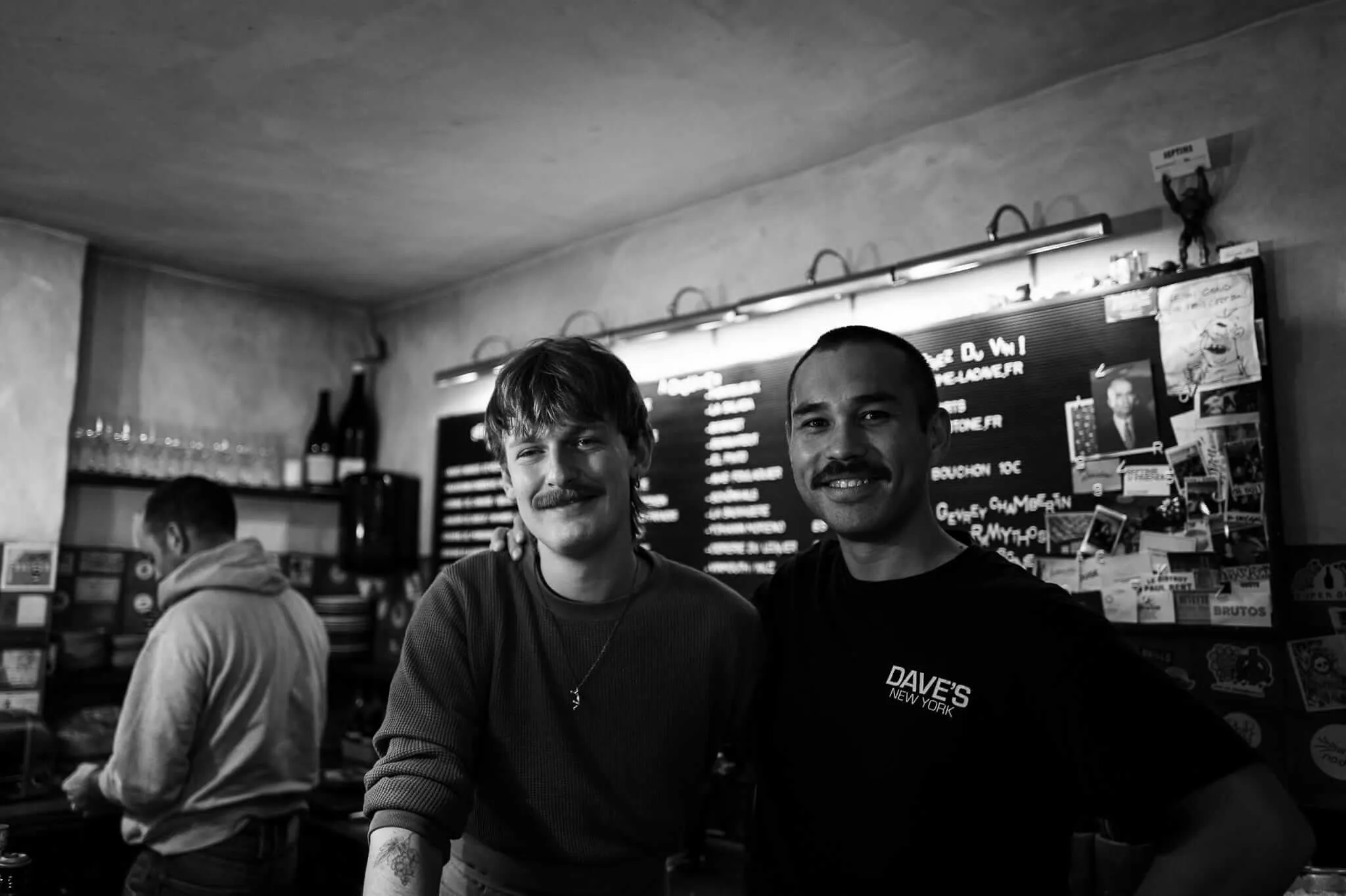 Black and white photograph of two smiling men with mustaches standing behind a counter in what appears to be a small restaurant or cafe, with menu boards visible in the background and another person partially visible to the left