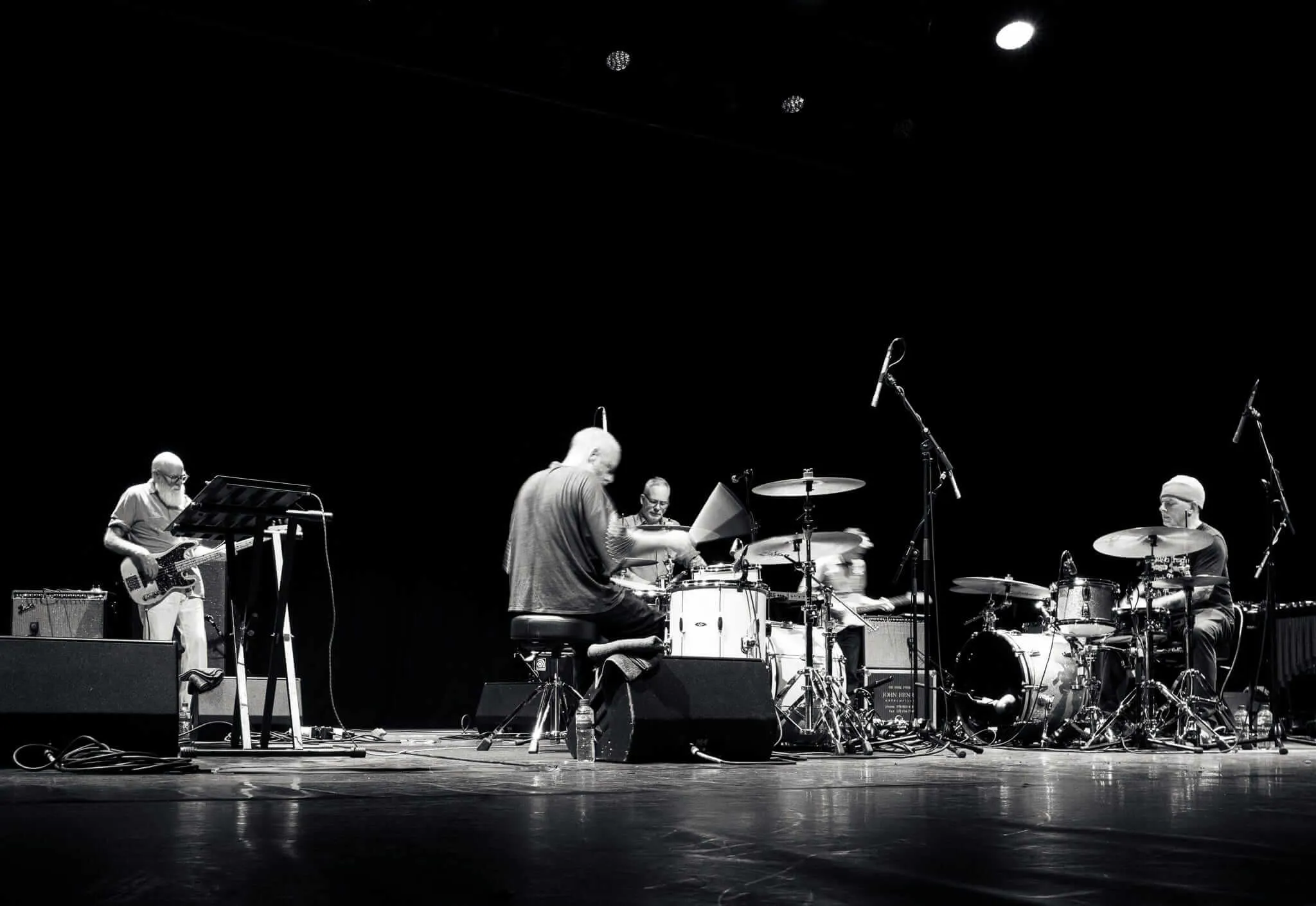 Black and white photograph of five jazz musicians performing on a dark stage, with a bassist on the left, drummer in center, and three other musicians on the right, all illuminated by stage lights against a black background