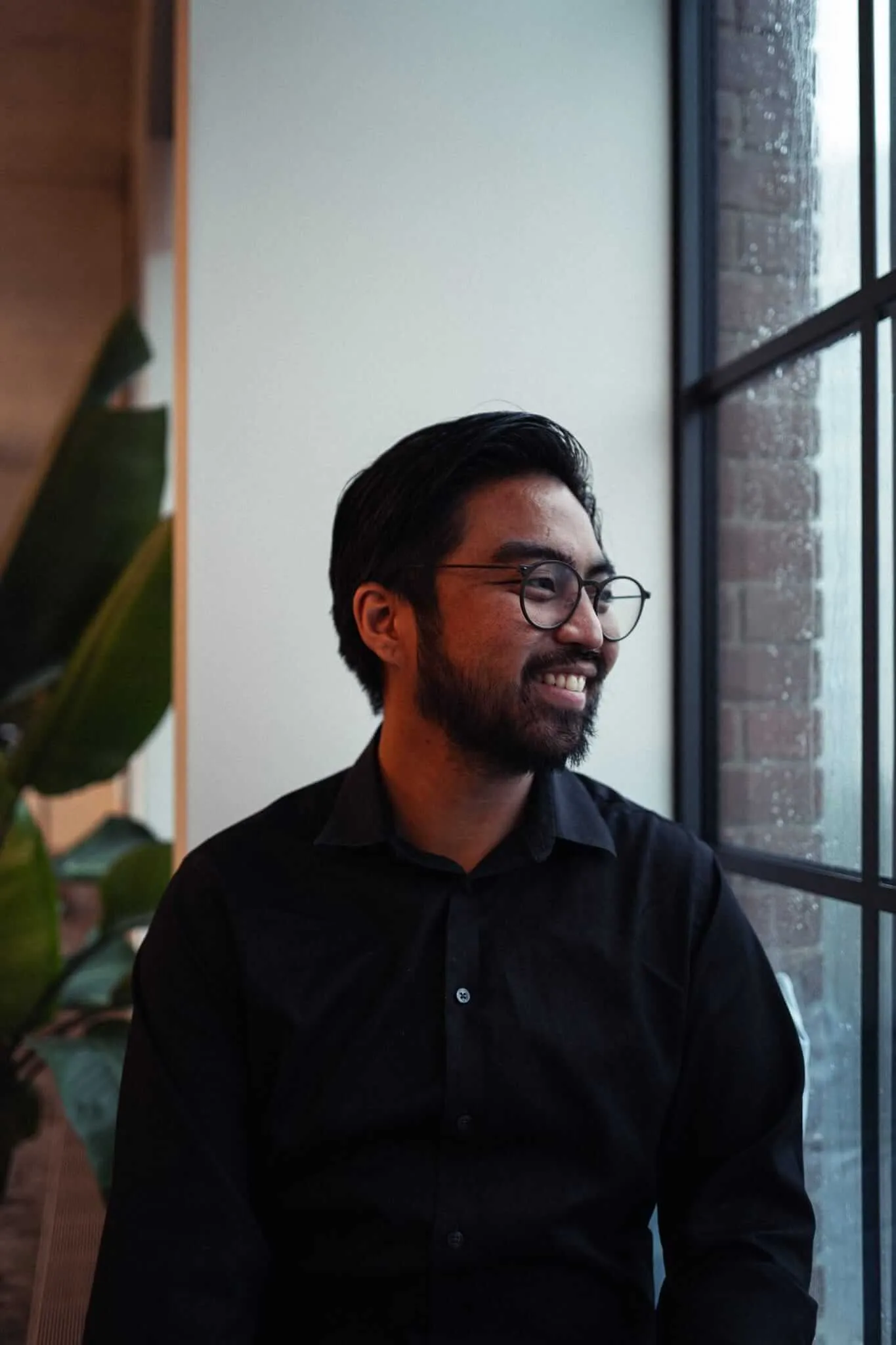 A young bearded man with round glasses and dark hair smiles warmly while gazing off to the side, seated near a rain-speckled industrial window beside a lush tropical plant, wearing a black button-up shirt in a softly lit interior space.