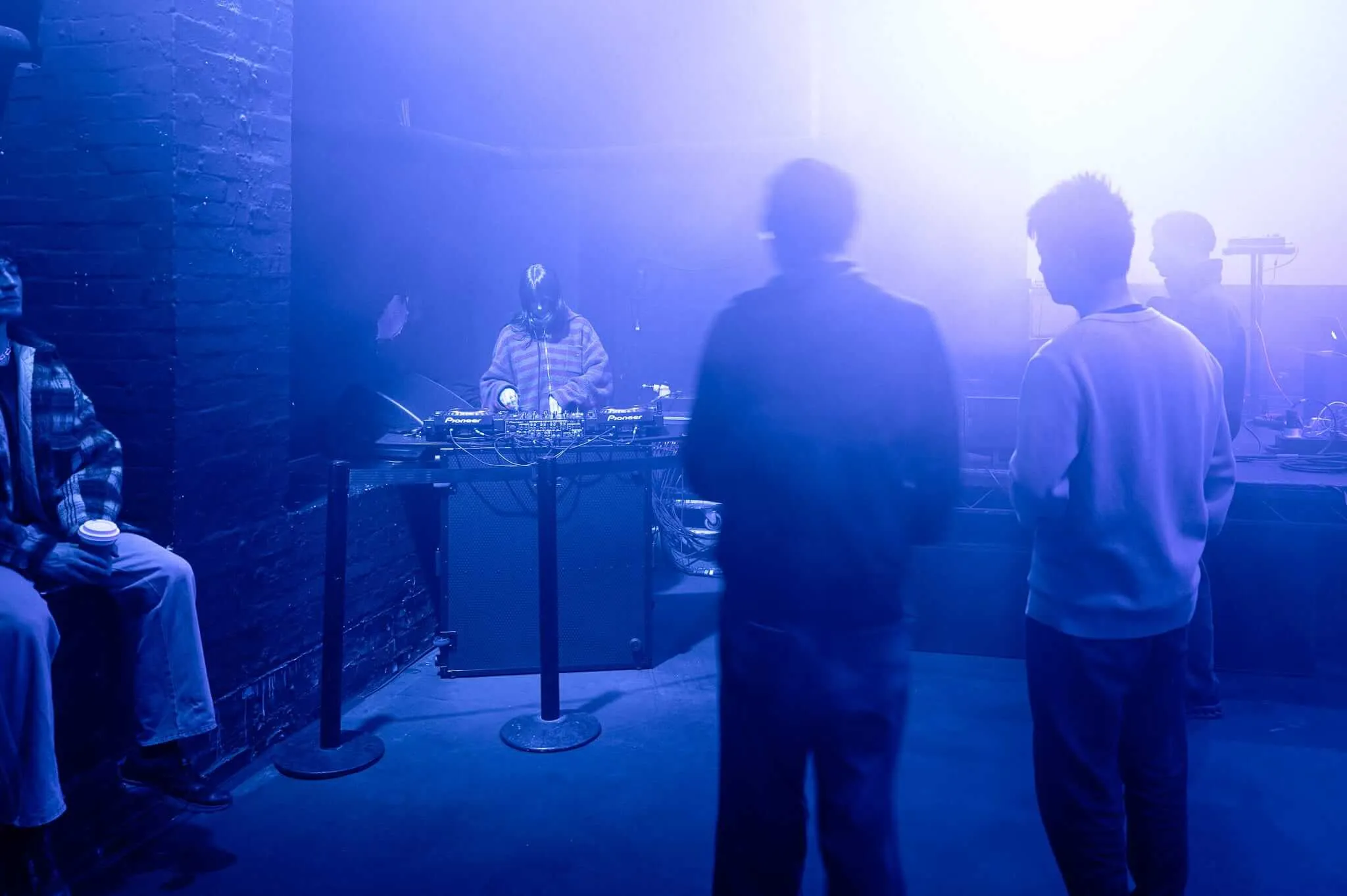 A DJ performs at a small venue bathed in intense blue and purple stage lighting, with several silhouetted audience members standing in the foreground watching the performance. The atmospheric lighting creates a moody, intimate club environment with dramatic contrasts between light and shadow.