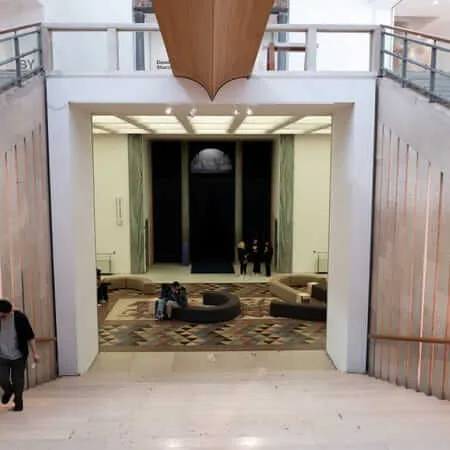 View from above looking down a wide stone staircase into a museum lobby featuring a geometric mosaic floor, curved modular seating, and visitors exploring an exhibition space with dramatic dark curtained installation beyond, flanked by copper-accented stone walls and upper gallery balconies.