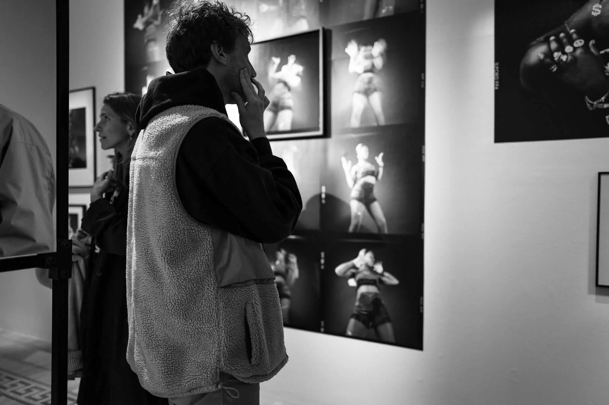 Black and white photograph of a person in a textured coat covering their face with their hand while viewing framed photographs in an art gallery. Multiple framed images are visible on the white walls behind them.