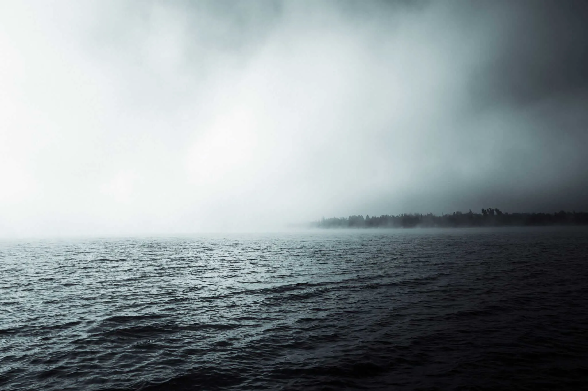 A dark, moody lake stretching toward a distant treeline barely visible through dense fog and low cloud cover, with gently rippling water in the foreground and a palette of deep grey-blue tones.