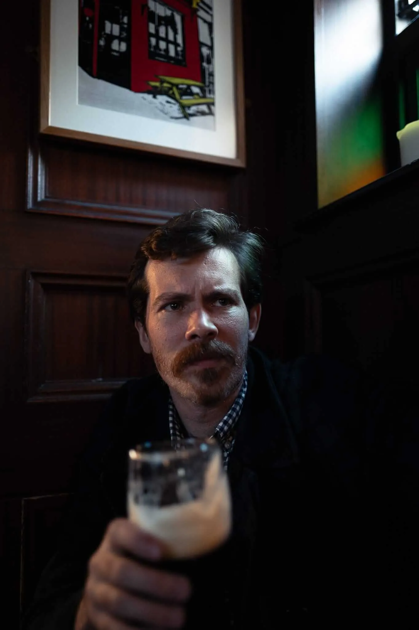 A contemplative man with a mustache sits in a dimly lit pub booth, holding a glass of beer. He wears a dark jacket over a checkered shirt, with warm lighting casting shadows across his face. Framed artwork hangs on dark wood paneling behind him.