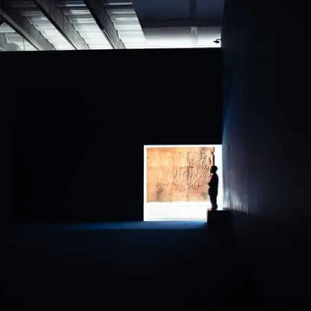 A lone silhouetted figure stands in a darkened museum gallery, gazing at a large illuminated artwork depicting a textured rock or earth surface, framed by a bright doorway at the end of a long, shadowy corridor.