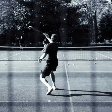 Black and white photograph of a tennis player mid-serve on an outdoor court, captured from behind as they reach up with their racket. The court is surrounded by trees and has tennis balls scattered across the surface, creating dramatic shadows in the natural lighting.