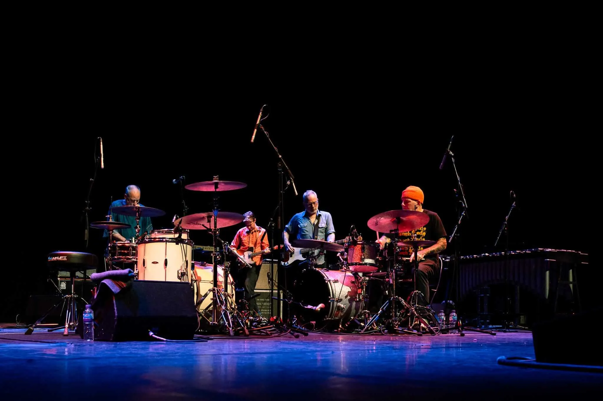 A band performing on a dark stage under colored stage lighting. From left to right: a performer working the electronics behind a white drum kit, a guitarist in an orange shirt, a bassist in a blue shirt, and a drummer in an orange beanie at a second drum set. The stage is illuminated with purple and pink lighting against a black background, with various microphone stands and equipment visible.