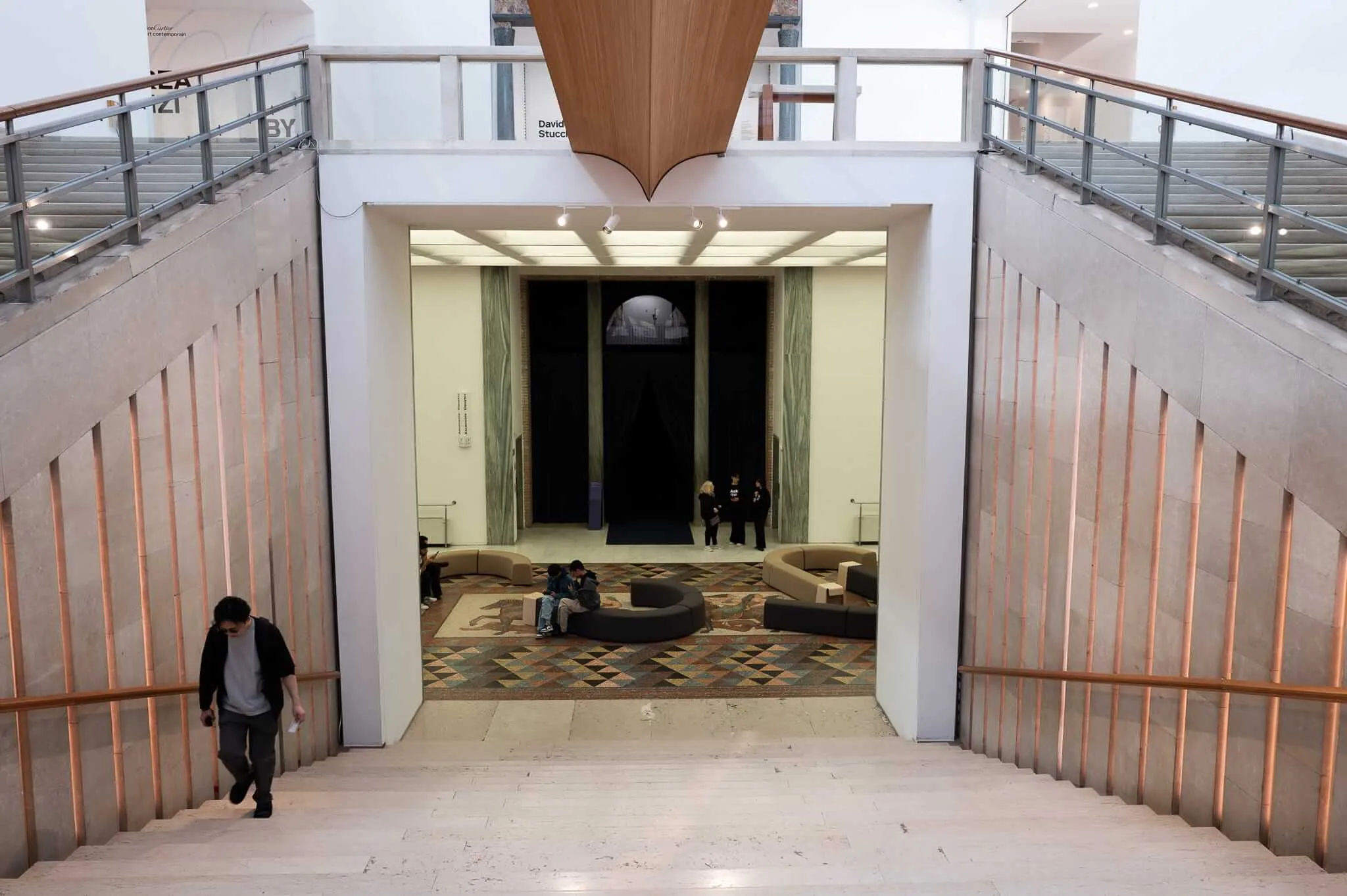 View from above looking down a wide stone staircase into a museum lobby featuring a geometric mosaic floor, curved modular seating, and visitors exploring an exhibition space with dramatic dark curtained installation beyond, flanked by copper-accented stone walls and upper gallery balconies.