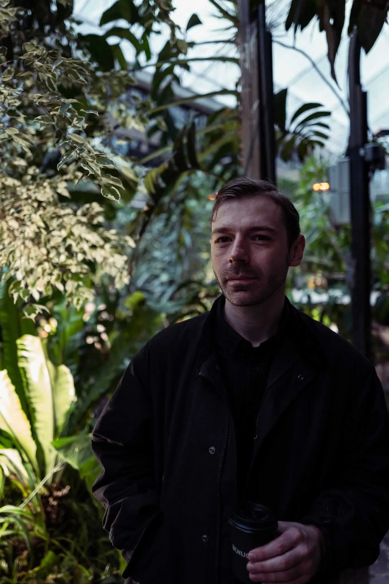 A man with dark hair and facial hair wearing a black shirt sits contemplatively in a greenhouse or conservatory setting, surrounded by lush tropical plants including palms and broad-leafed foliage, with natural light filtering through glass panels in the background