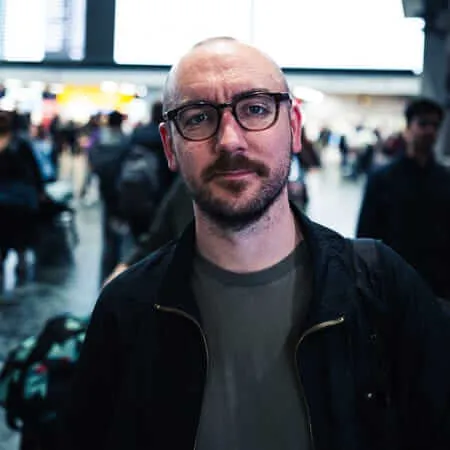 A man with black-rimmed glasses and a mustache wearing a dark jacket and gray t-shirt stands in the foreground of what appears to be an airport terminal, with bright overhead lighting and blurred crowds of travelers in the background