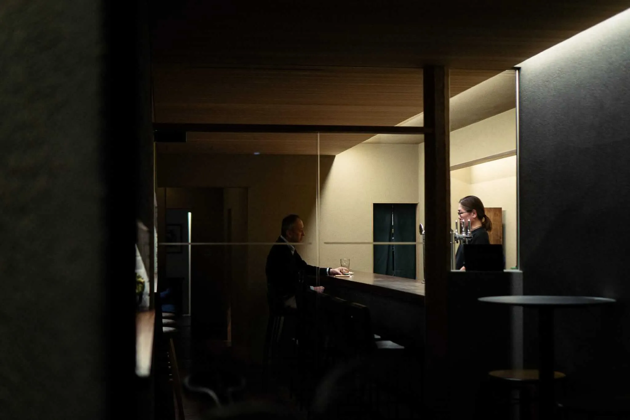 A dimly lit scene viewed through a doorway or window frame, showing a man in a dark suit seated at a bar counter holding a glass, while a woman wearing glasses stands behind the counter in conversation with him, bathed in warm ambient light against a minimal Japanese-inspired interior.