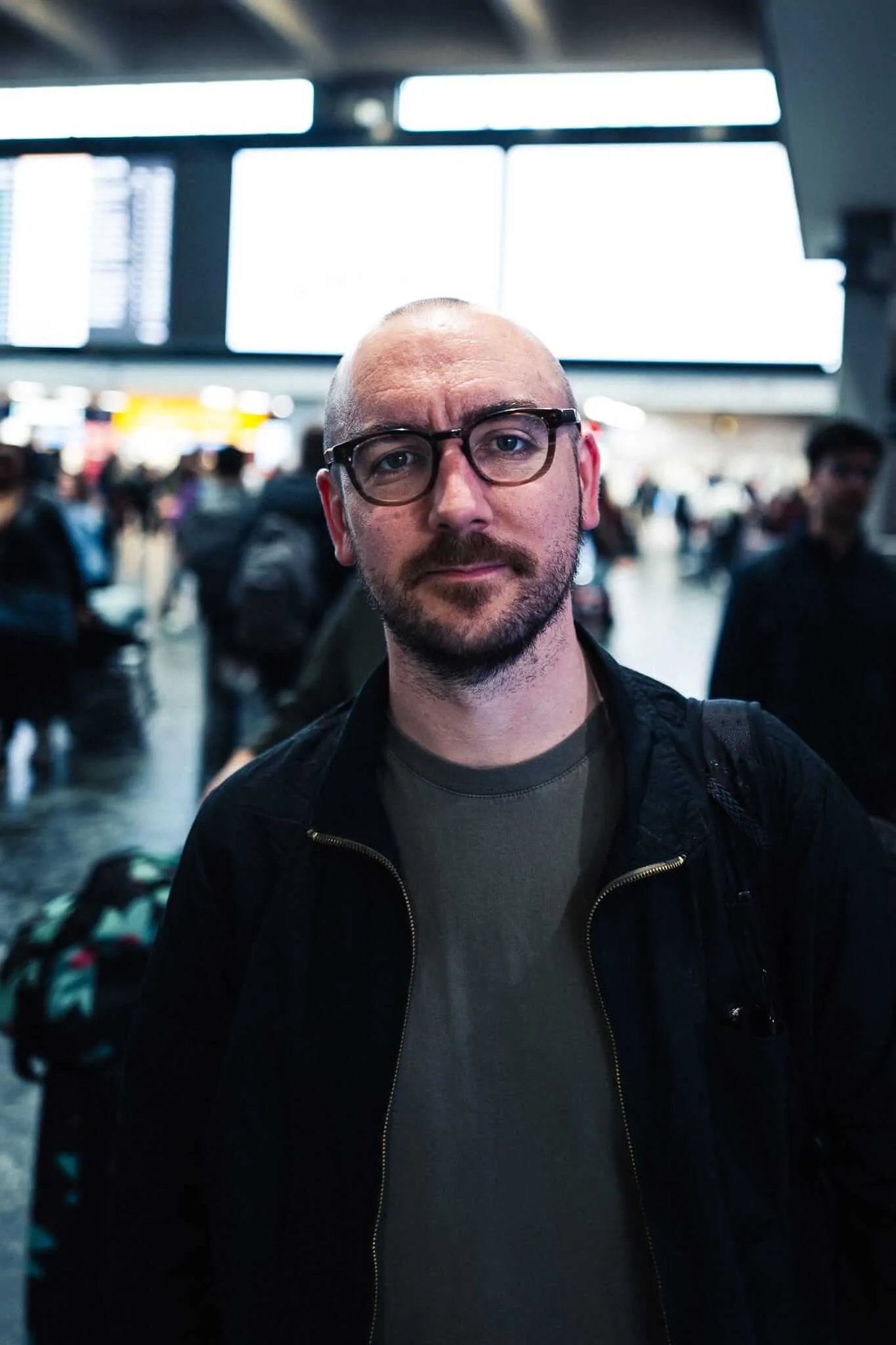 A man with black-rimmed glasses and a mustache wearing a dark jacket and gray t-shirt stands in the foreground of what appears to be an airport terminal, with bright overhead lighting and blurred crowds of travelers in the background