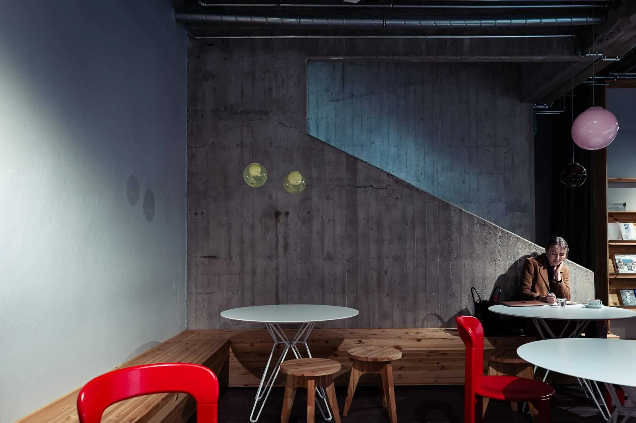 A modern interior space with exposed concrete walls and industrial ceiling elements. A person in a brown coat sits alone at a white round table in the background, reading or writing. The foreground features wooden bench seating with red plastic chairs and small white round tables. Colorful spherical light fixtures - yellow and pink orbs - hang from the ceiling, creating warm accent lighting against the brutalist architecture.