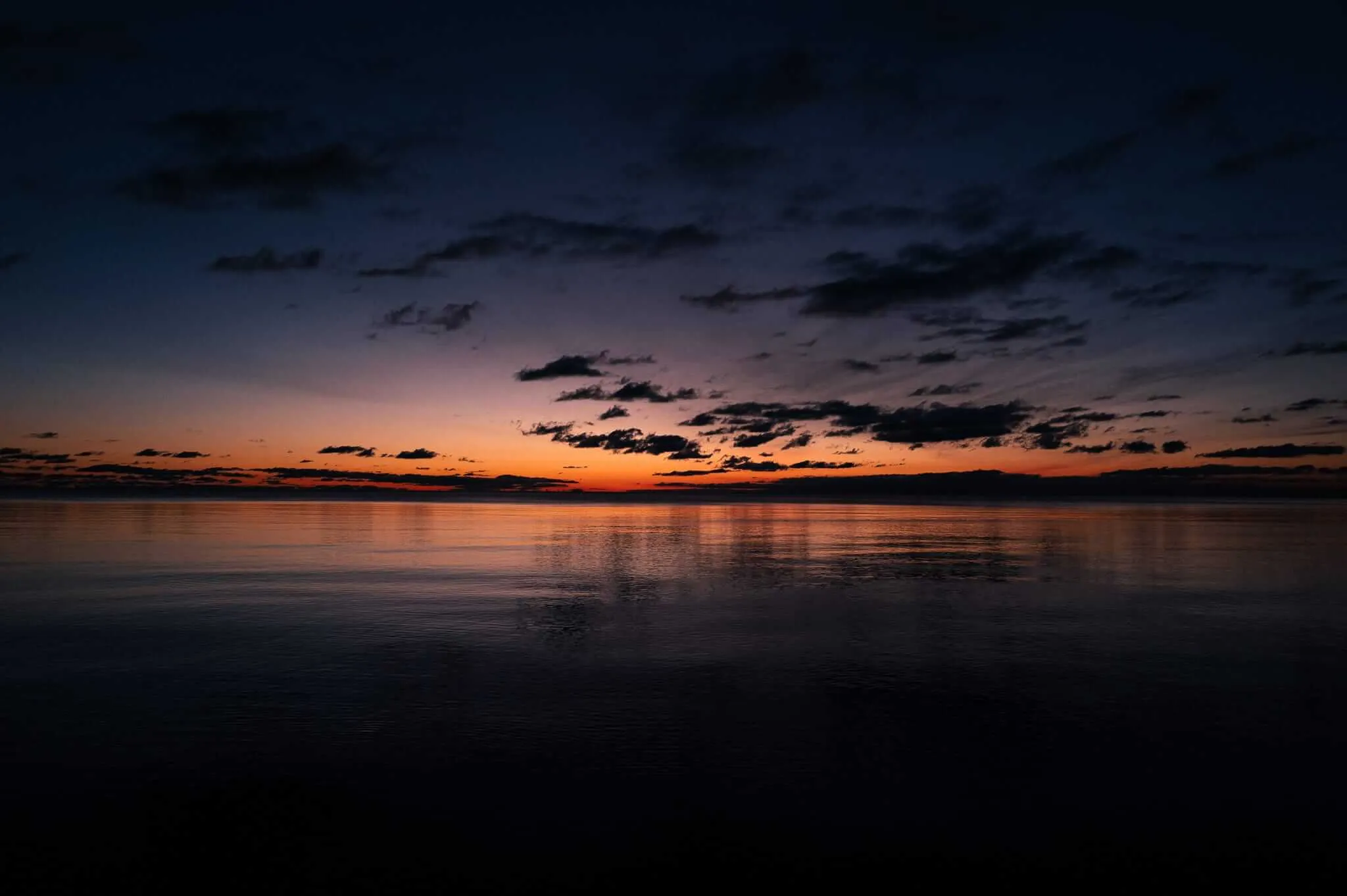 A serene twilight seascape showing a perfectly calm body of water reflecting dramatic clouds and a vibrant orange sunset glow on the horizon, creating a mirror-like surface that doubles the sky's beauty