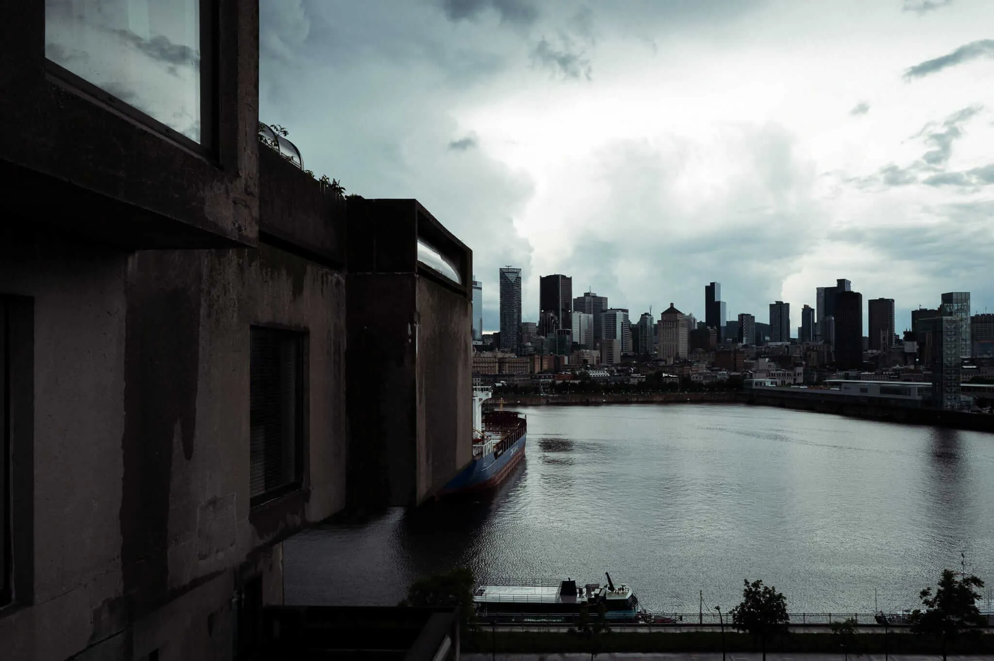 A moody, dark cityscape viewed from beside a weathered concrete building, with a cargo ship navigating a wide river in the foreground and Montreal's downtown skyline stretching across the horizon under a dramatically overcast sky.