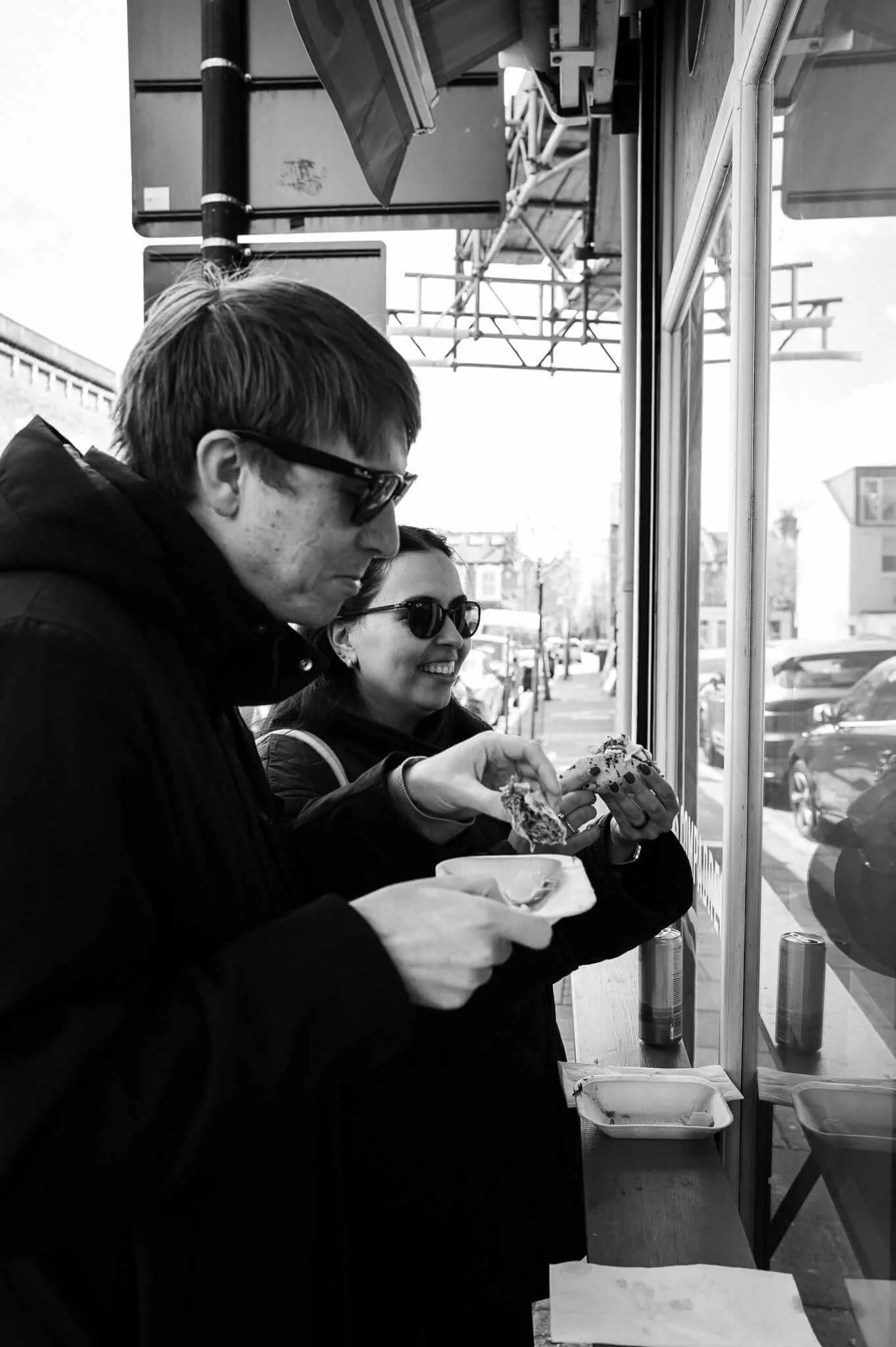 Black and white photograph of a couple at a restaurant counter by large windows. A man in glasses stands behind a woman who is also wearing sunglasses and smiling. She holds what appears to be a taco. The scene is lit by natural daylight streaming through the windows, with urban street views visible outside.