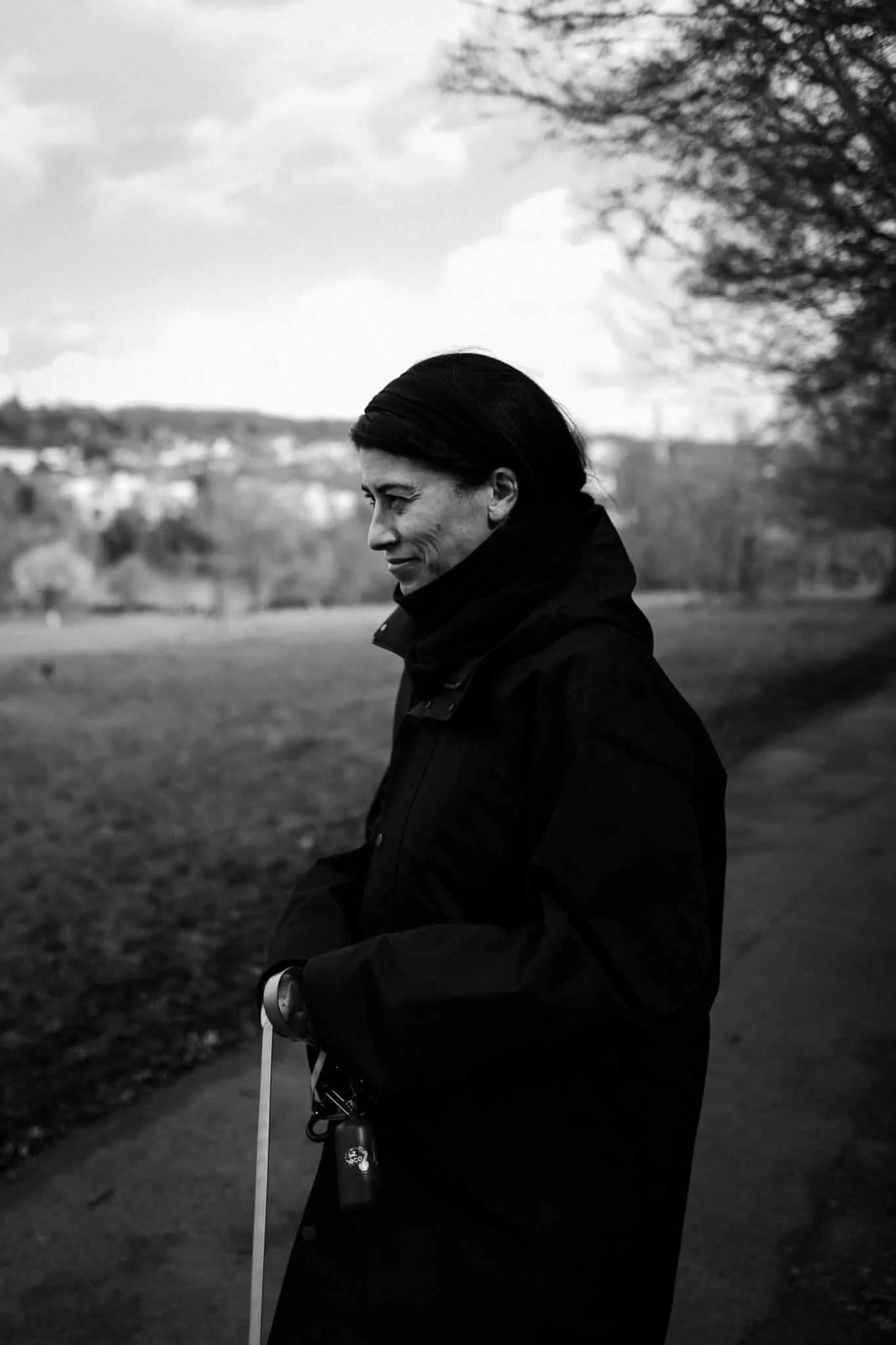 A black and white portrait of a woman in profile wearing a dark coat, holding a dog lead, standing in an open field with trees and buildings blurred in the background under an overcast sky