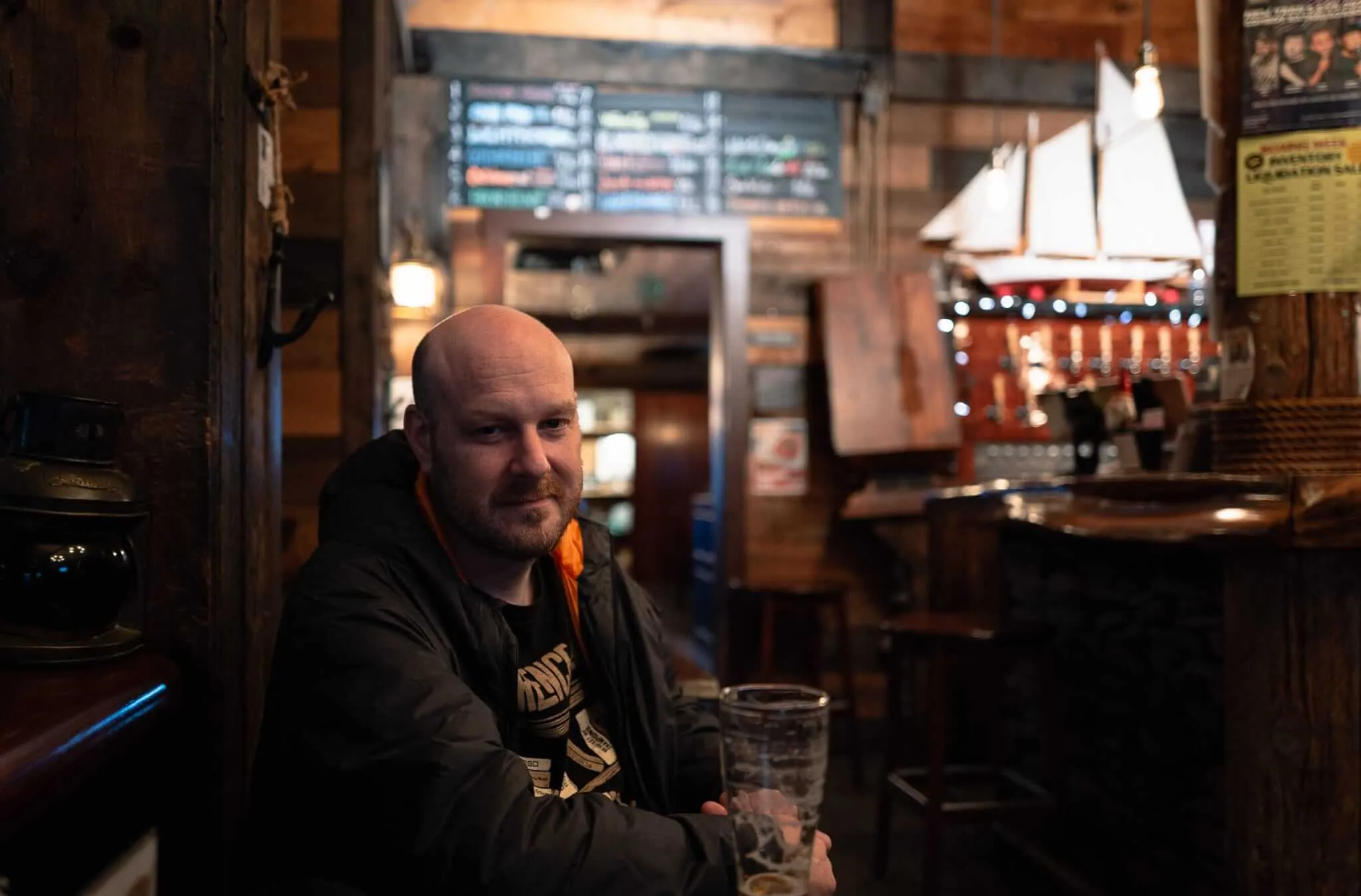 A man with a beard sits at a wooden table in a dimly lit nautical-themed bar, smiling warmly at the camera. Behind him, a model sailboat with white sails hangs from the ceiling, and wooden walls display maritime memorabilia including what appears to be a menu board. The atmosphere is cozy and intimate with warm amber lighting.