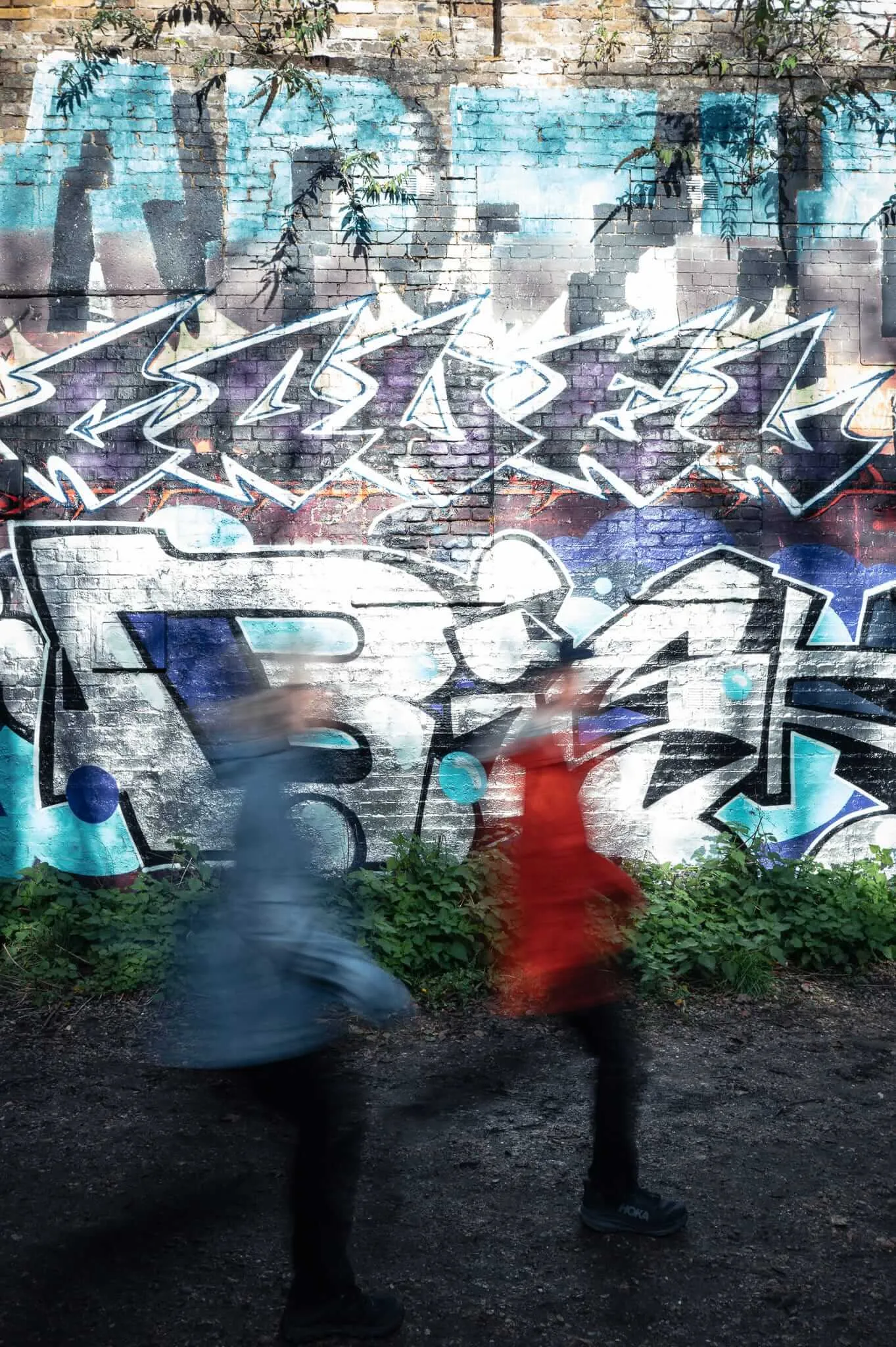 Motion-blurred figures in blue and red clothing walk past a brick wall covered in colorful graffiti, with angular white lettering above and stylized text below, creating streaks of movement against the static urban art backdrop