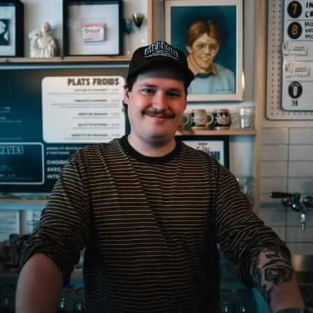 A young male bartender with a mustache, tattoos, and a black 'Mellon' baseball cap smiles warmly behind a bar counter. The eclectic wall behind him features framed artwork including a painted portrait, shelves with decorative objects, a 'Plats Froids' menu board, and a numbered list, suggesting a French-language establishment.
