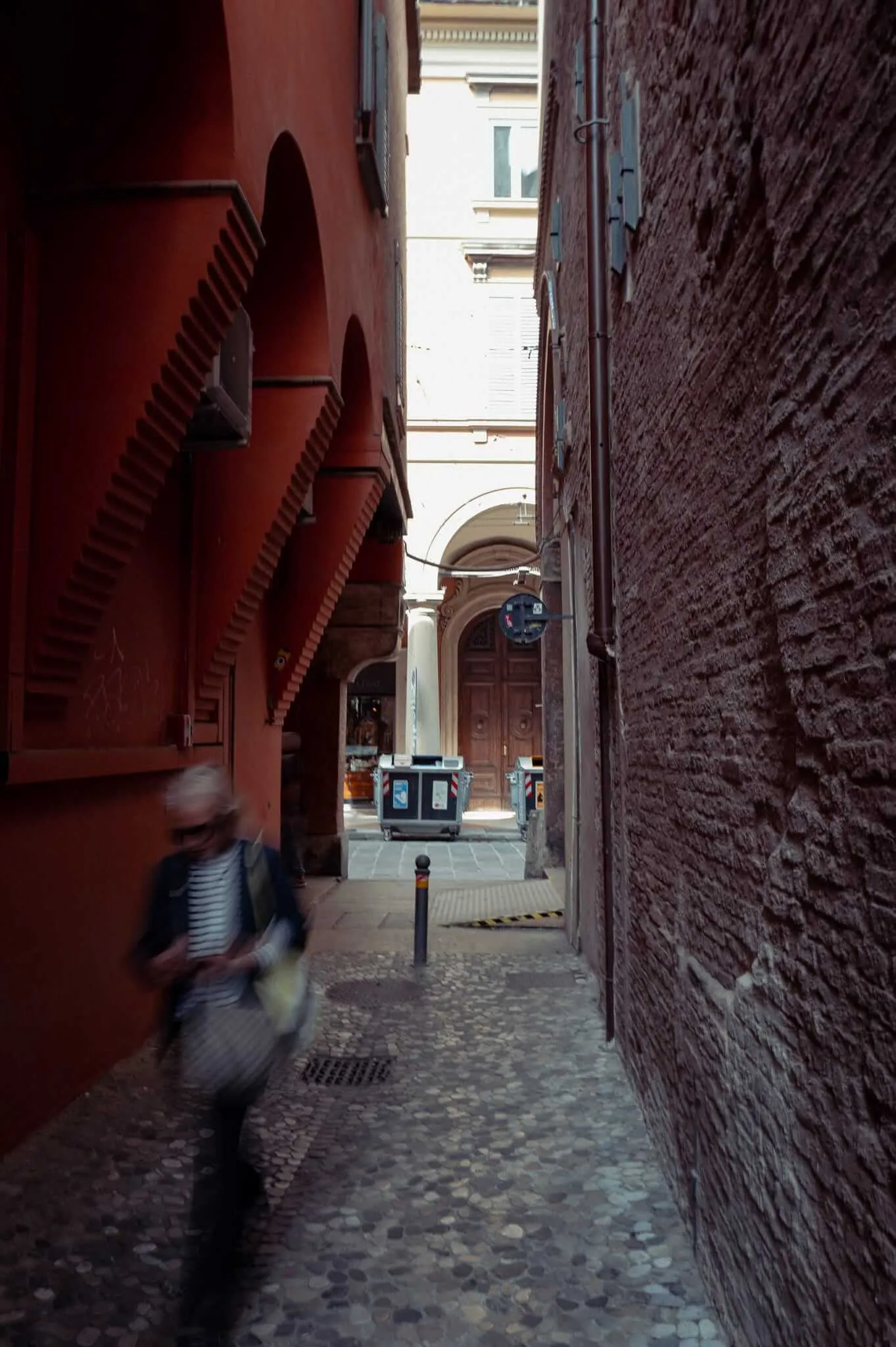 A narrow cobblestone alley in an Italian city with warm terracotta and brick walls. A person in a striped shirt walks quickly through the passage, creating motion blur. The alley opens to a brighter square with classical architecture and arched doorways visible in the background.