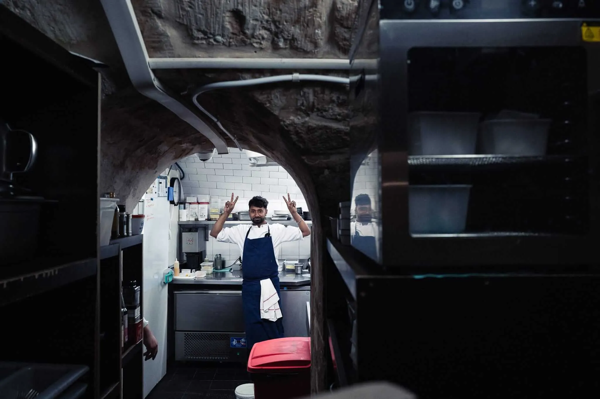 A chef in white jacket and blue apron stands in a professional kitchen making peace signs with both hands, framed by a stone archway. The kitchen has stainless steel equipment, white tile walls, and industrial fixtures visible in the background.