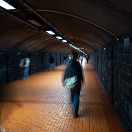 A long exposure photograph taken inside a pedestrian tunnel with an arched brick ceiling and warm orange tiled floor. Motion-blurred figures walk through the dimly lit underground passage, illuminated by a row of fluorescent ceiling lights receding into the distance. The closest figure carries a bag and appears ghost-like due to the long shutter speed.