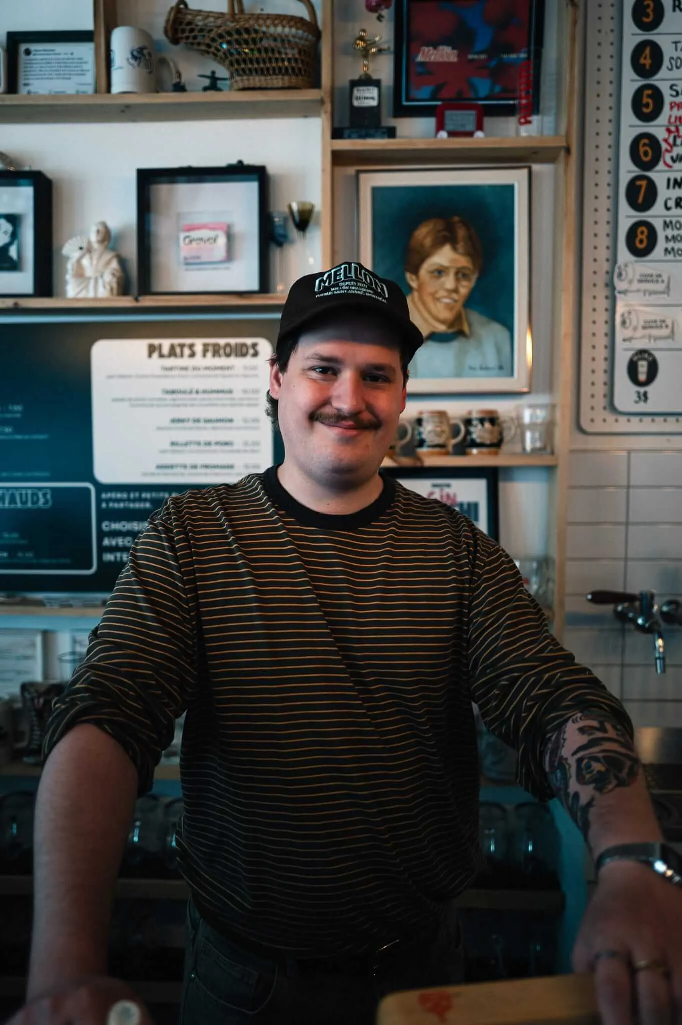 A young male bartender with a mustache, tattoos, and a black 'Mellon' baseball cap smiles warmly behind a bar counter. The eclectic wall behind him features framed artwork including a painted portrait, shelves with decorative objects, a 'Plats Froids' menu board, and a numbered list, suggesting a French-language establishment.