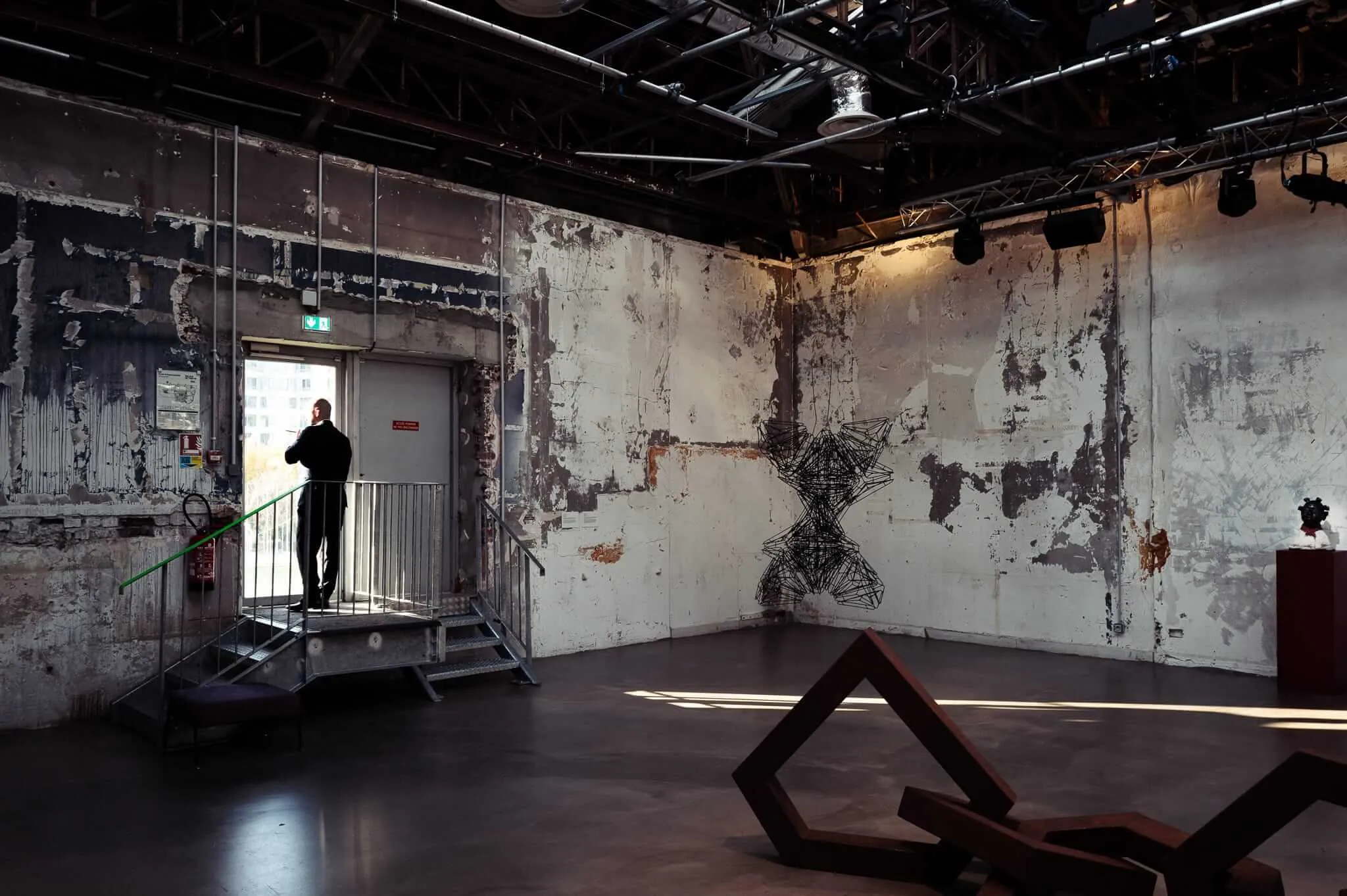 A person in dark clothing stands on metal stairs in a deteriorating industrial gallery space with peeling white walls, exposed ceiling infrastructure, and abstract wire sculptures mounted on the walls. The scene combines decay with contemporary art installation.