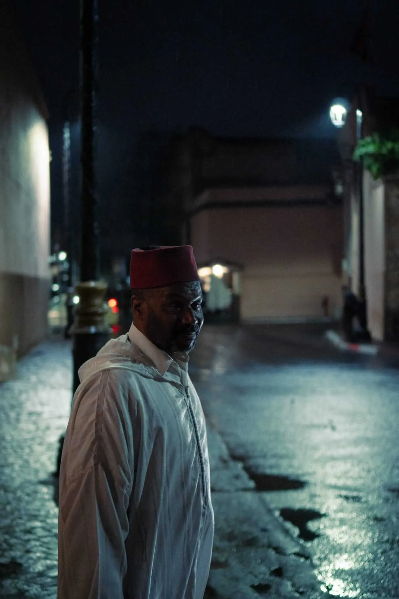 A man wearing a traditional red fez and white djellaba stands on a rain-slicked cobblestone alleyway at night, glancing back over his shoulder toward the camera, with dim streetlights illuminating the passage behind him.