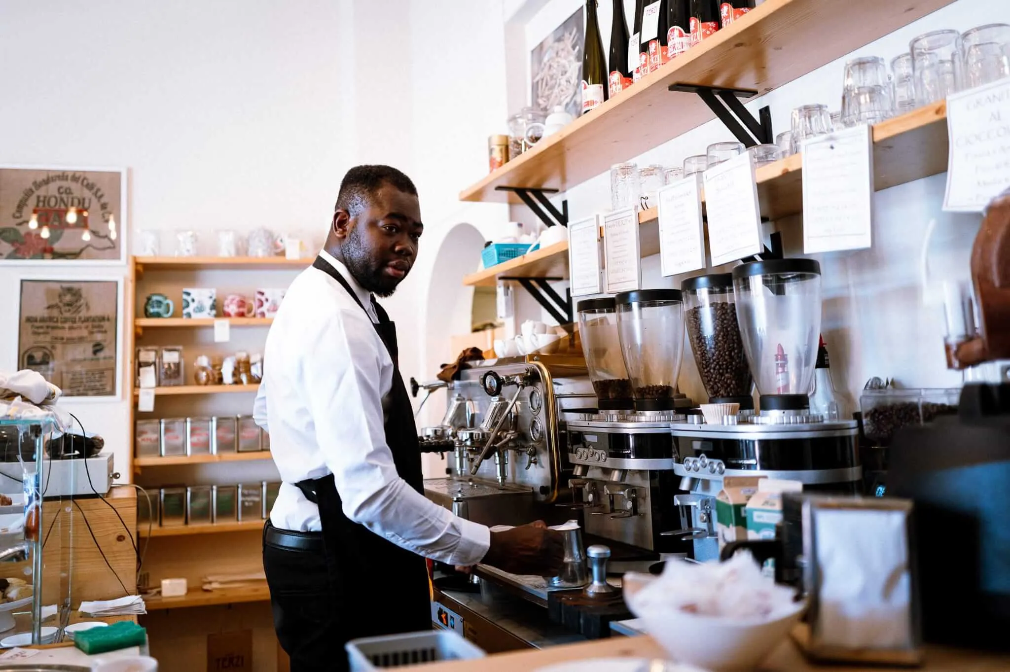 A barista in white shirt and dark apron working behind an espresso machine in a modern coffee shop, with wooden shelves displaying bottles and glasses, coffee grinders visible, and warm lighting throughout the space