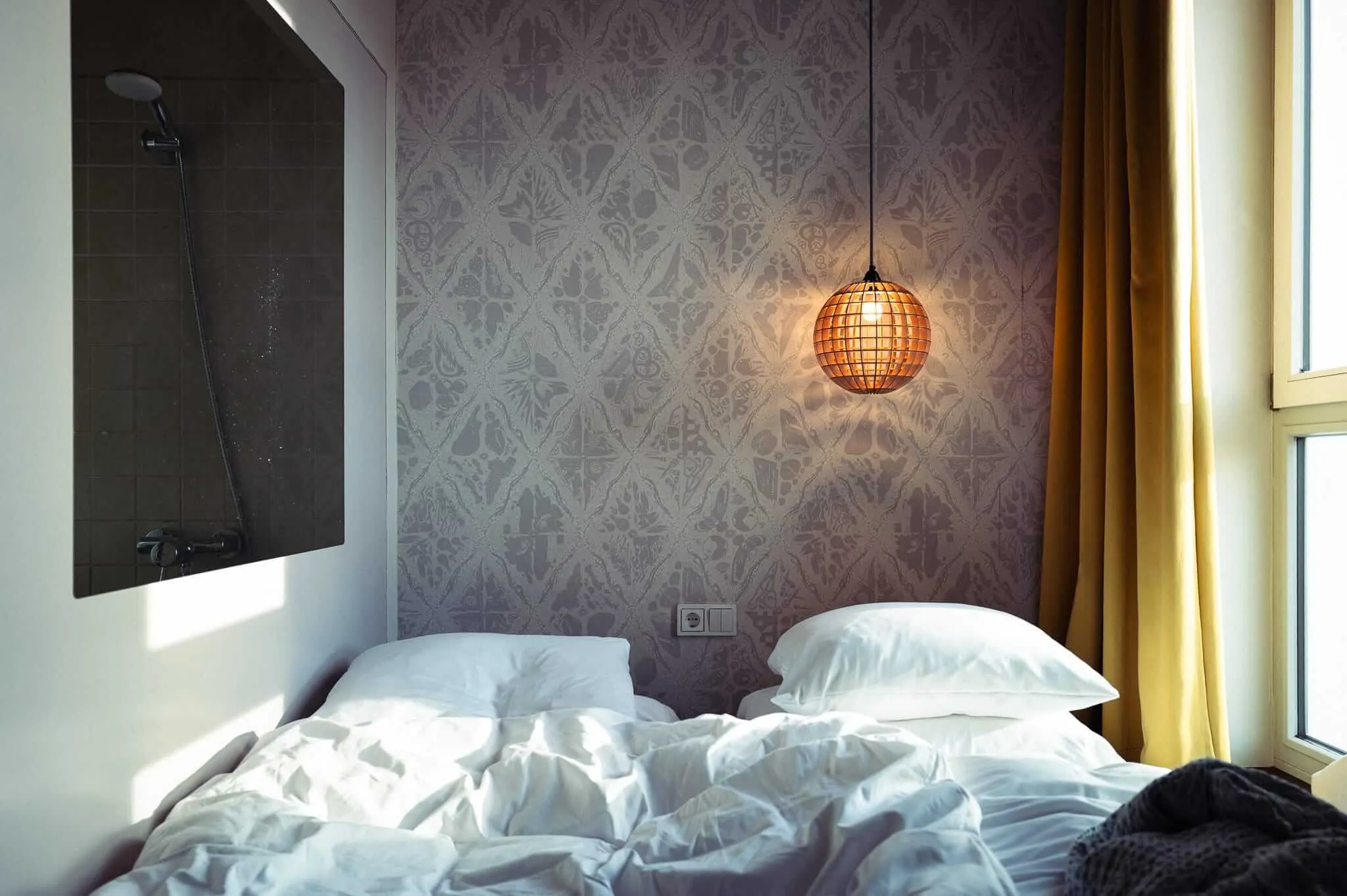A hotel bedroom with an unmade bed featuring white bedding, a decorative patterned wallpaper headboard, and a distinctive spherical woven pendant light casting warm light. Golden curtains frame a window on the right, and a view of the shower on the left wall.