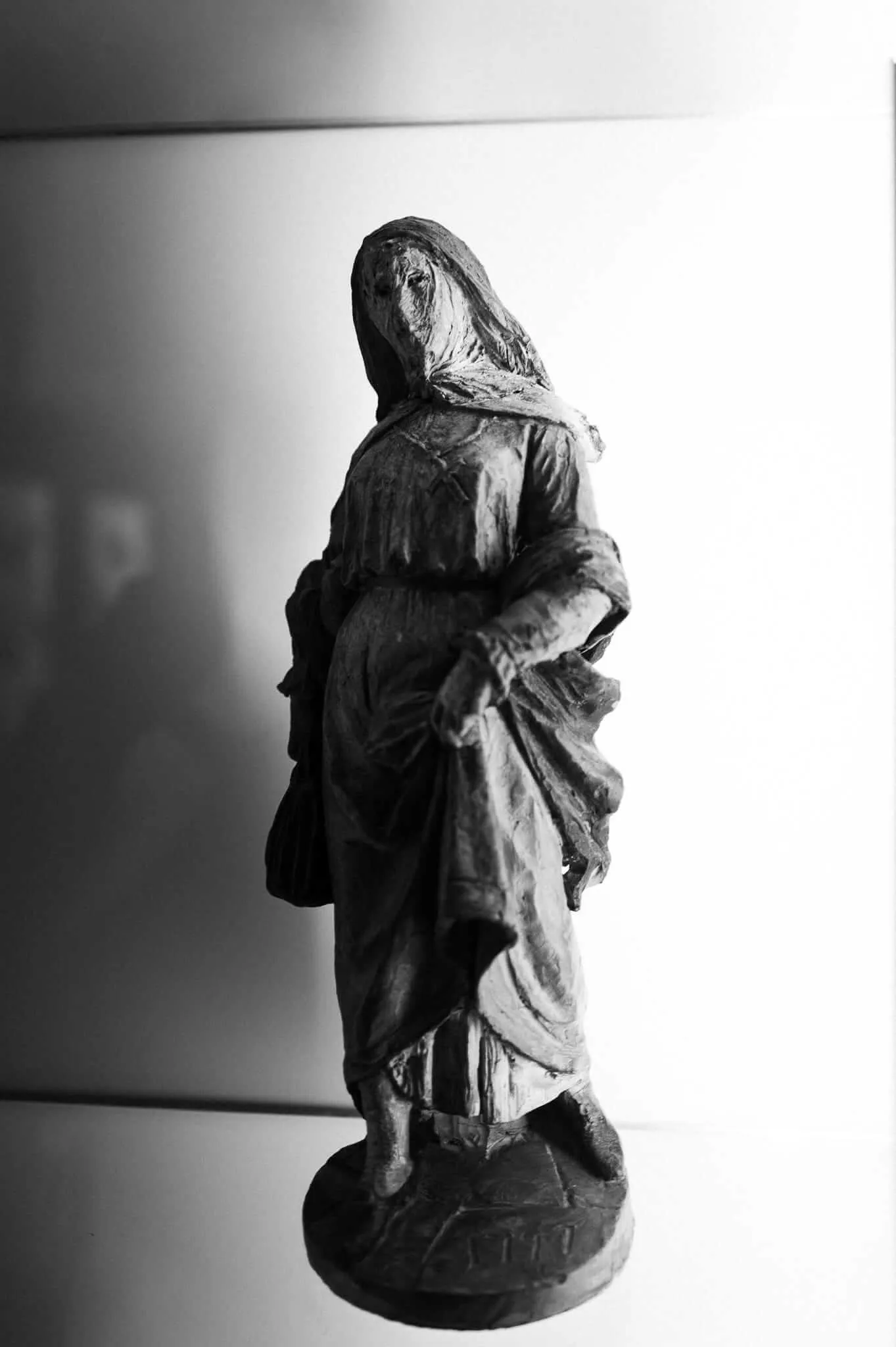 A dark, weathered sculpture of a robed and hooded female figure standing on a circular base, photographed in black and white with dramatic chiaroscuro lighting against a bright white background.
