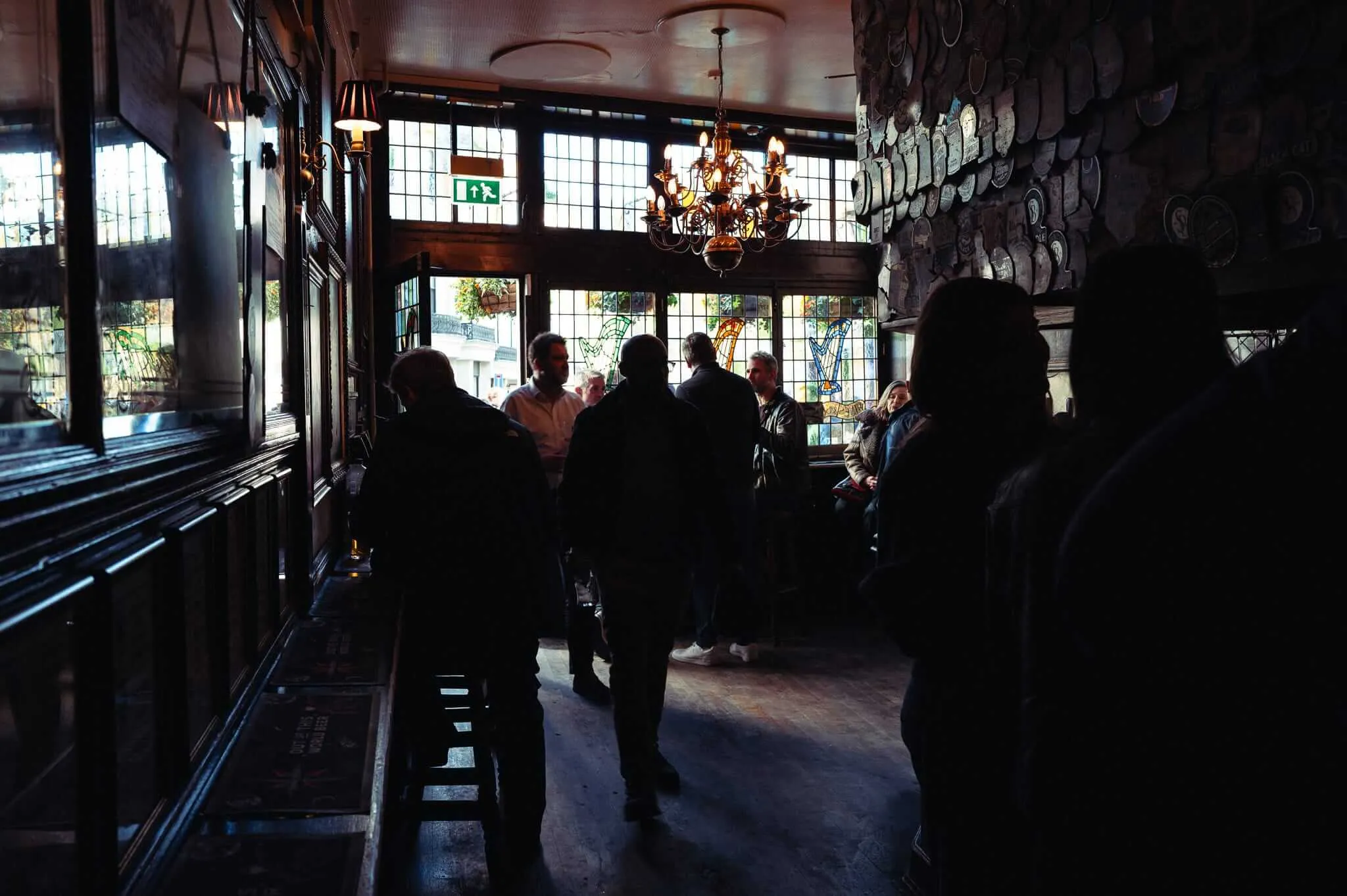 Interior of a traditional pub with dark wood paneling, ornate chandelier, and plates displayed on walls. Several patrons are silhouetted against bright windows with stained glass elements, creating a moody atmosphere with strong contrast between light and shadow.