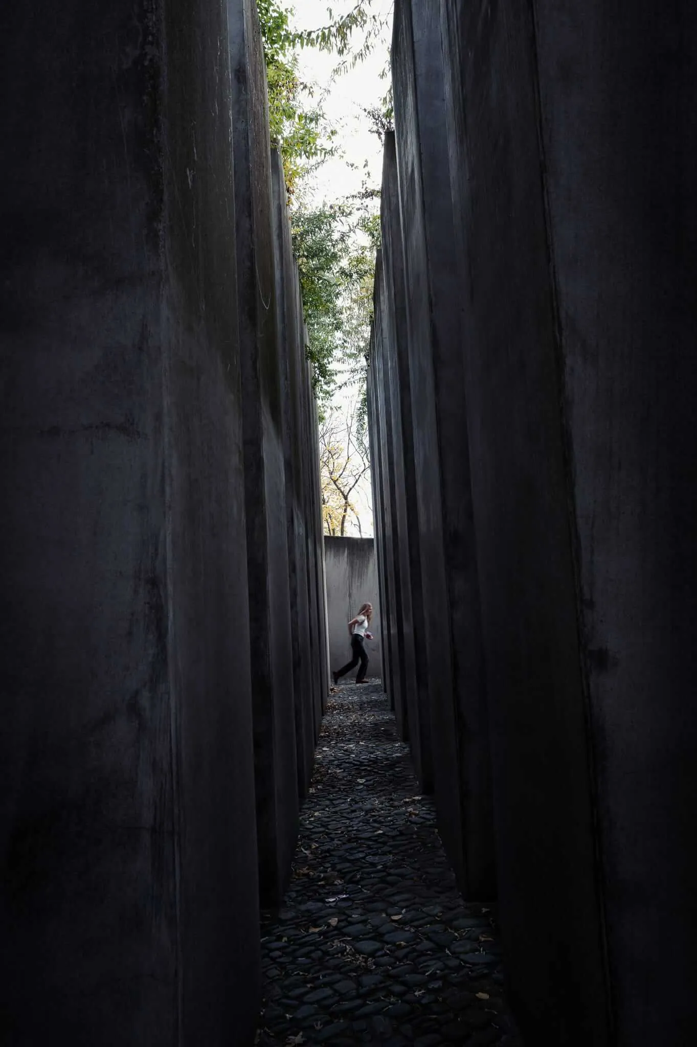A narrow passageway between tall concrete slabs or monuments, with a small figure of a person walking through the shadowy corridor. The path appears to be paved with cobblestones, and green foliage is visible at the top opening where natural light filters down.