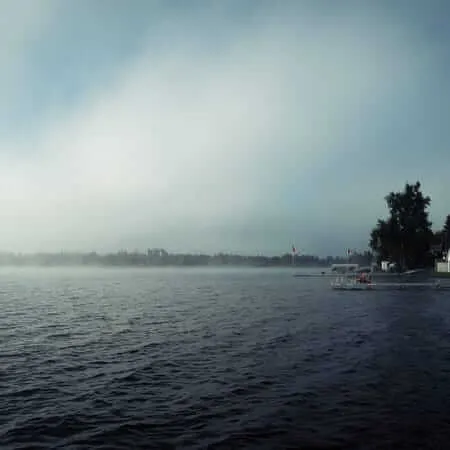 A misty morning over a dark, rippling lake with fog rolling across the far treeline. On the right shore, docks, cottages, and Canadian flags are partially visible among dense trees under an overcast sky.