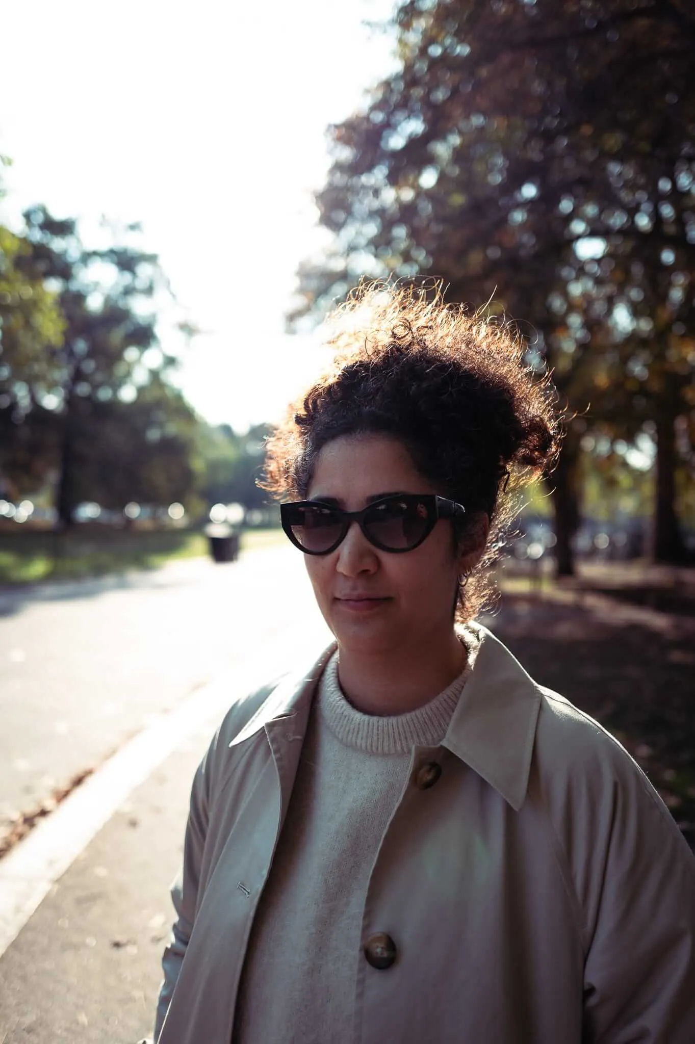 A woman with curly dark hair wearing sunglasses, a cream sweater, and beige trench coat stands on a tree-lined street. Golden backlighting creates a luminous halo effect around her hair as she faces the camera with a slight smile.