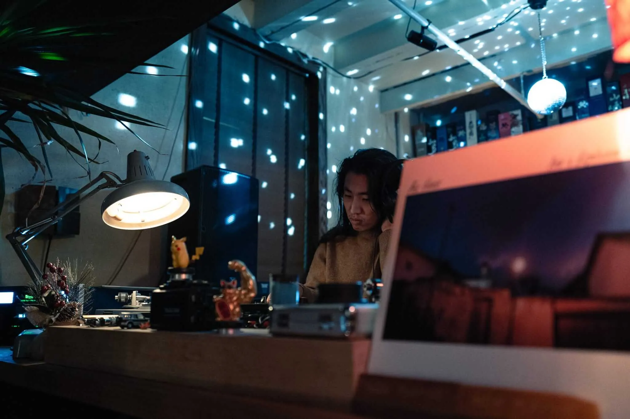 A person with dark hair wearing a brown sweater stands behind dj decks in a dimly lit modern interior space. The scene is illuminated by a desk lamp and ambient lighting, with bokeh light effects creating a constellation-like pattern across the walls and ceiling. The workspace appears to contain various objects and equipment, creating an atmospheric, contemplative setting.
