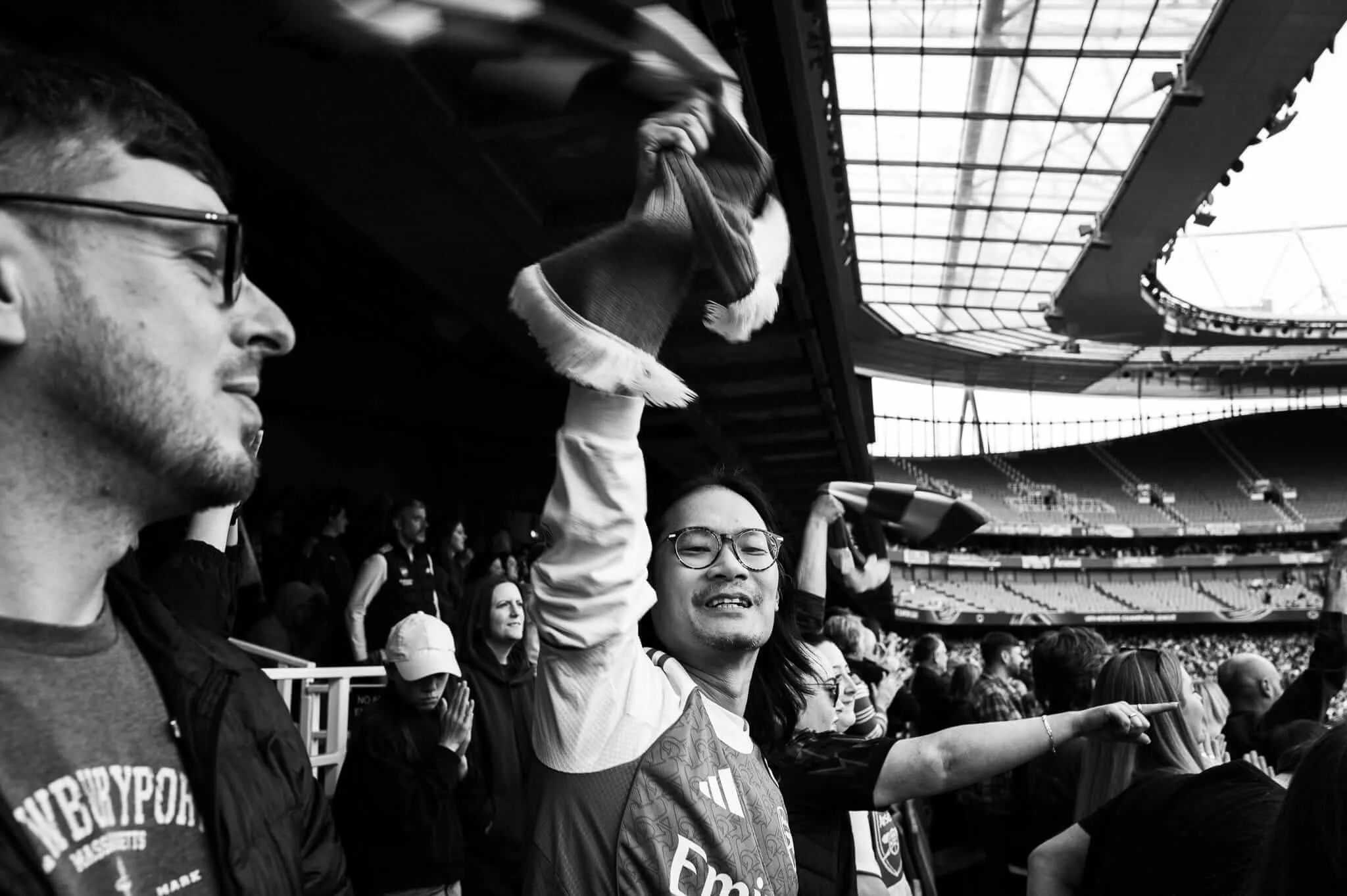 Black and white photograph of excited sports fans in a stadium, with a man in glasses and scarf celebrating with raised hands while another fan in a cap looks on, stadium architecture visible in the background