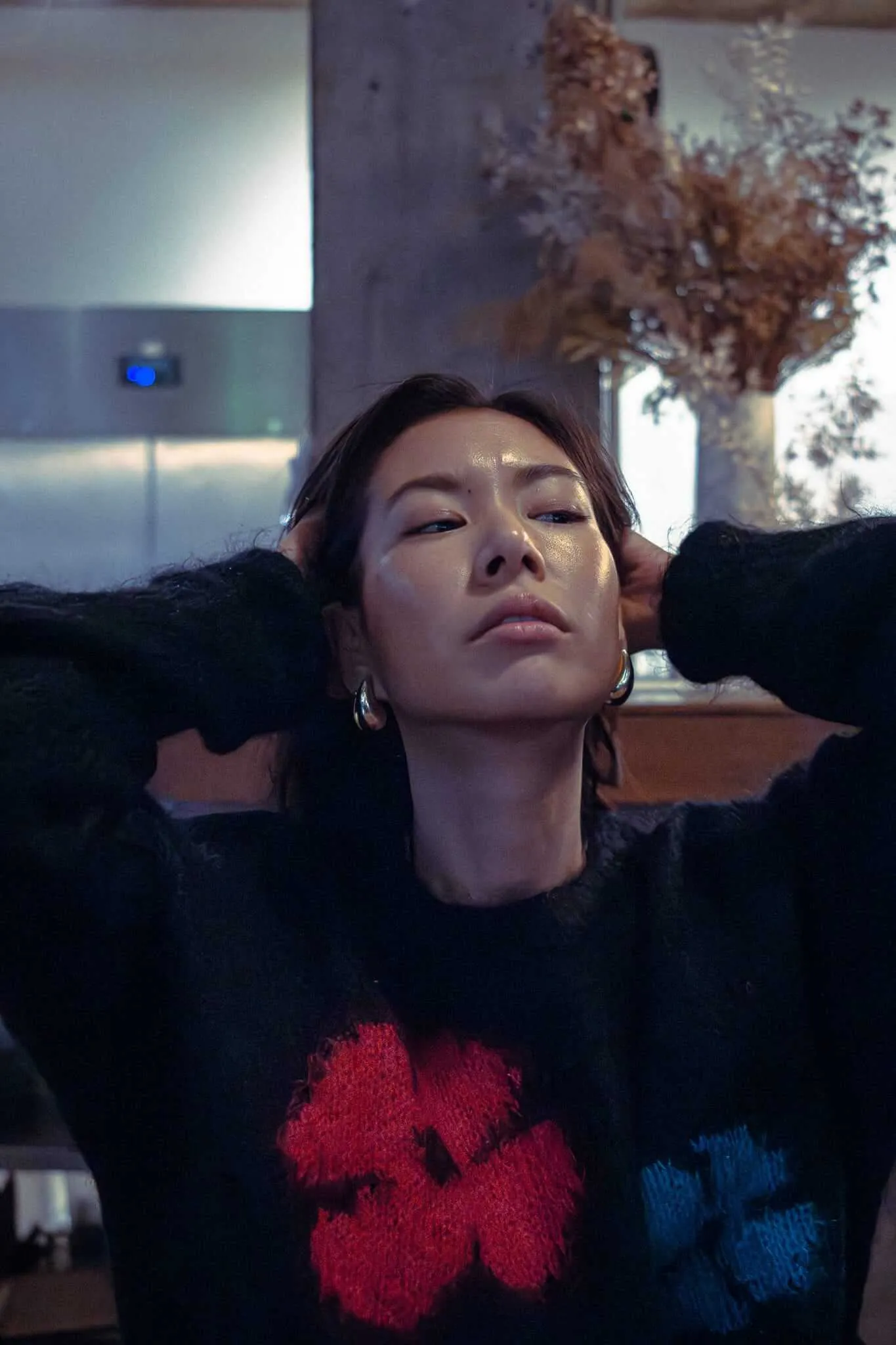 A woman with dark hair pulled back, eyes half-closed, wearing a black fuzzy sweater with red and blue floral patterns, her hands raised behind her head, posed against a softly lit interior background with dried floral arrangements.